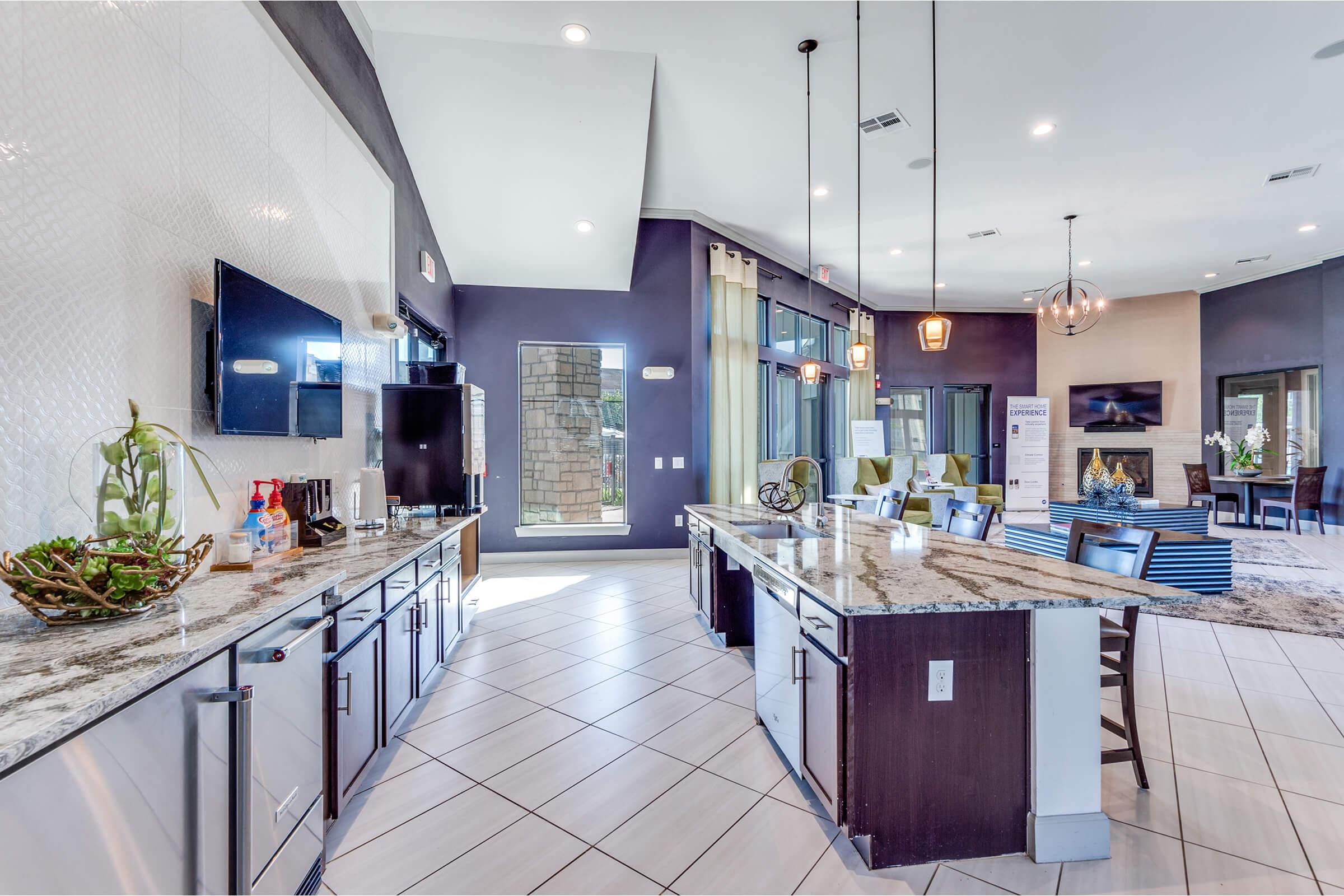 A modern kitchen area featuring a granite countertop with bar seating, sleek cabinetry, and appliances. Large windows allow natural light to flood in, highlighting the stylish interior design. The space also includes a lounge area with comfortable seating and decorative elements, creating a welcoming ambiance.