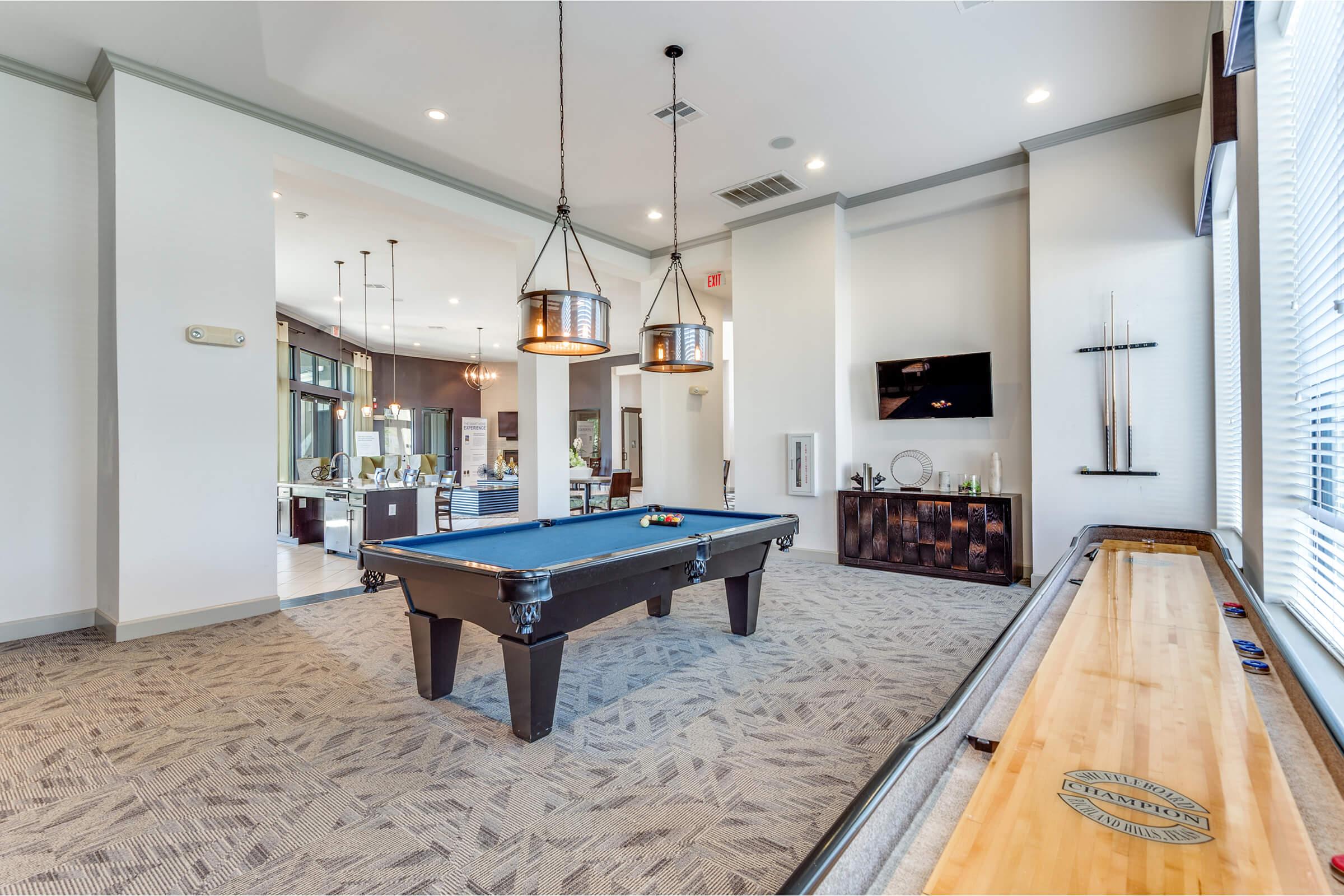 A modern recreational room featuring a billiards table with colorful balls on it. Two stylish pendant lights hang above the table, and windows provide natural light. In the background, there is a kitchen area with bar seating and a large TV mounted on the wall, alongside decorative shelves.