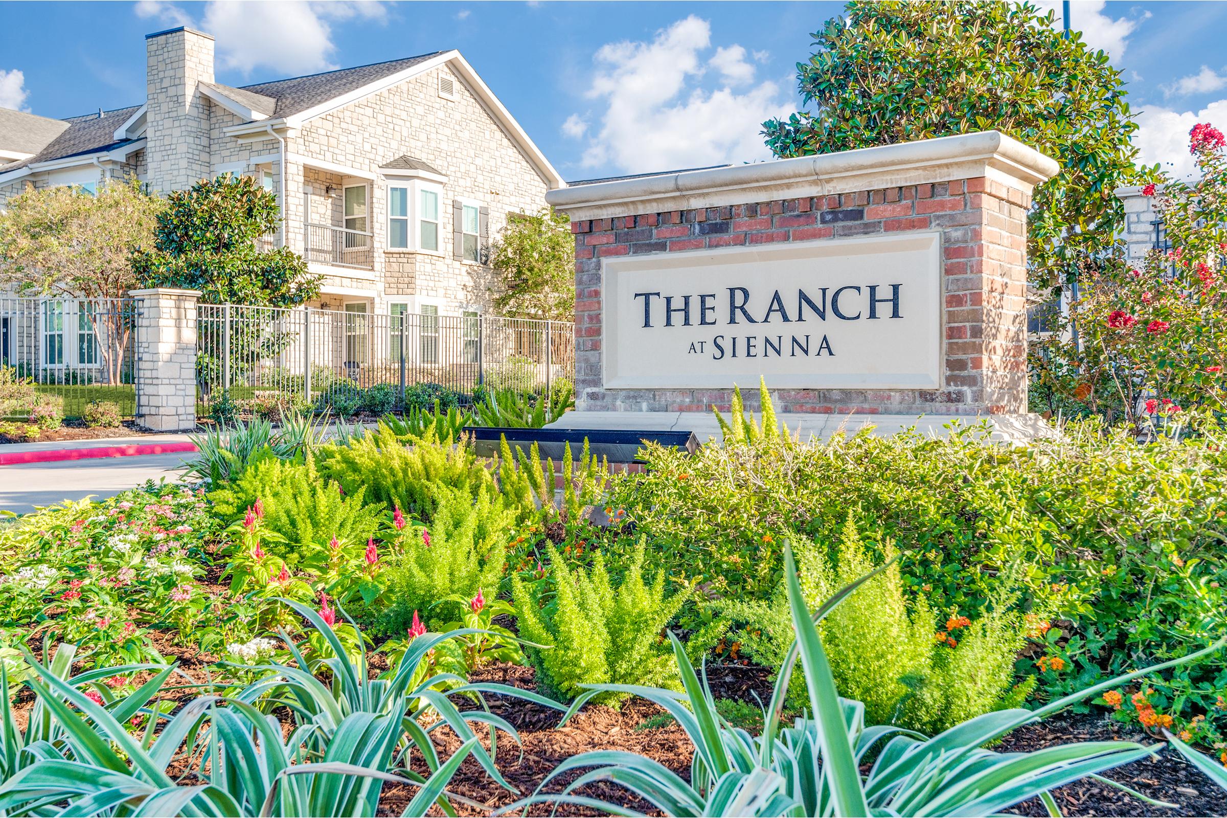 A welcoming sign for "The Ranch at Sienna" surrounded by vibrant landscaping, including various green plants and colorful flowers. In the background, there are residential buildings with stone facades under a clear blue sky.