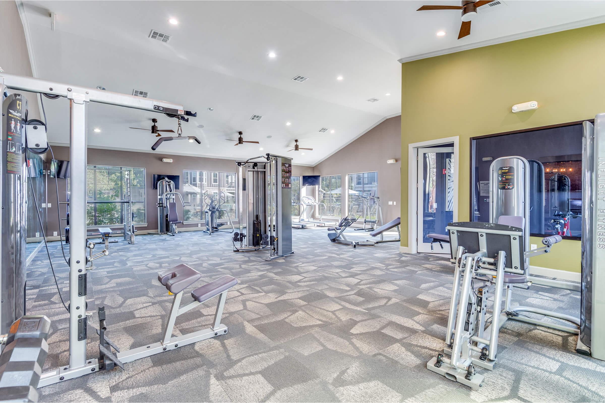 A spacious, well-lit gym area featuring various exercise machines, including weightlifting equipment and cardio machines. Large windows allow natural light, and the ceiling is high with ceiling fans. The walls are painted in light colors, creating a welcoming workout environment.
