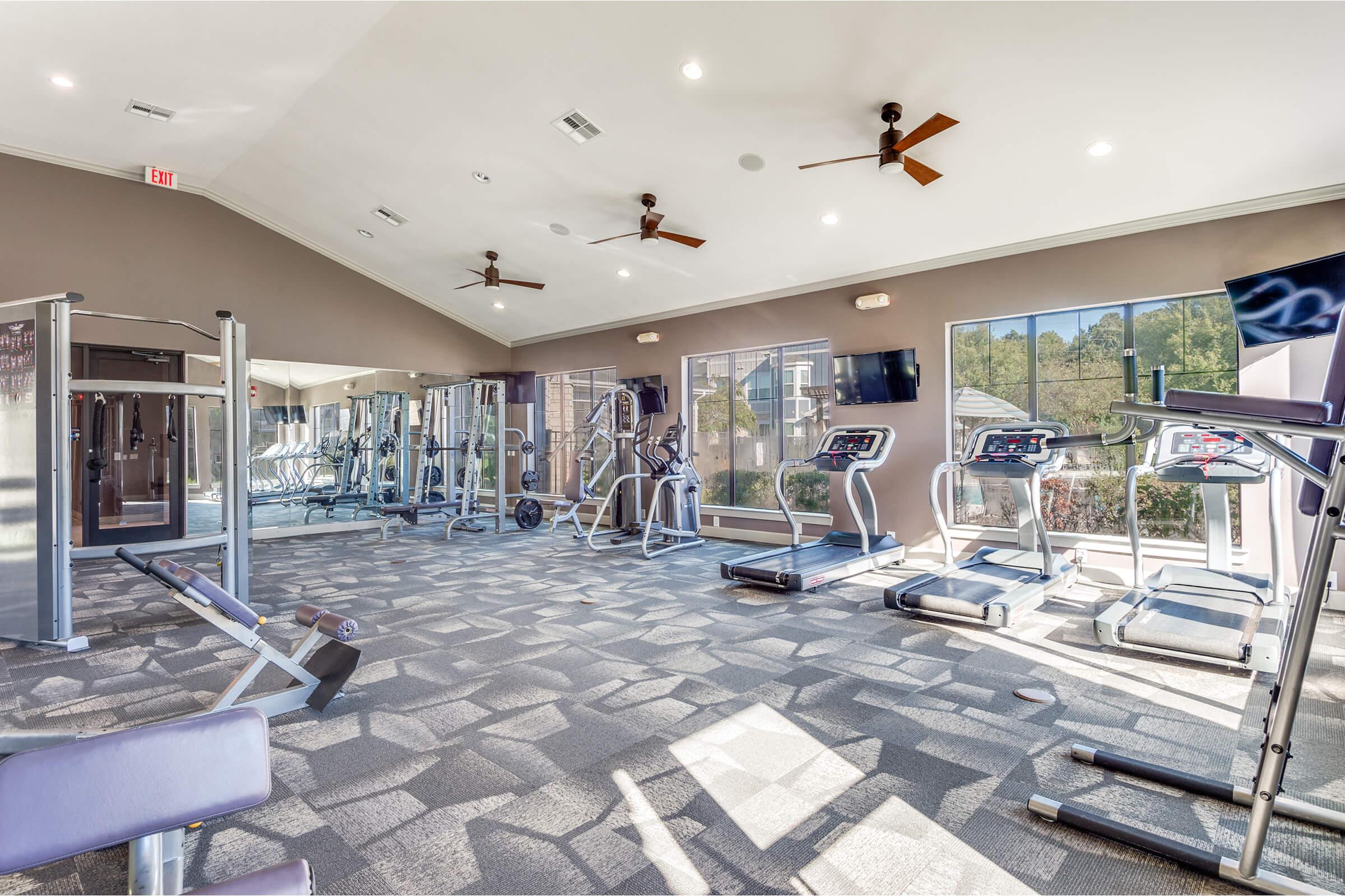 A modern gym interior featuring various exercise equipment, including treadmills, weight machines, and free weights. Large windows allow natural light to fill the space, and ceiling fans are installed for ventilation. The floor is covered with gray tiles, enhancing the contemporary design.