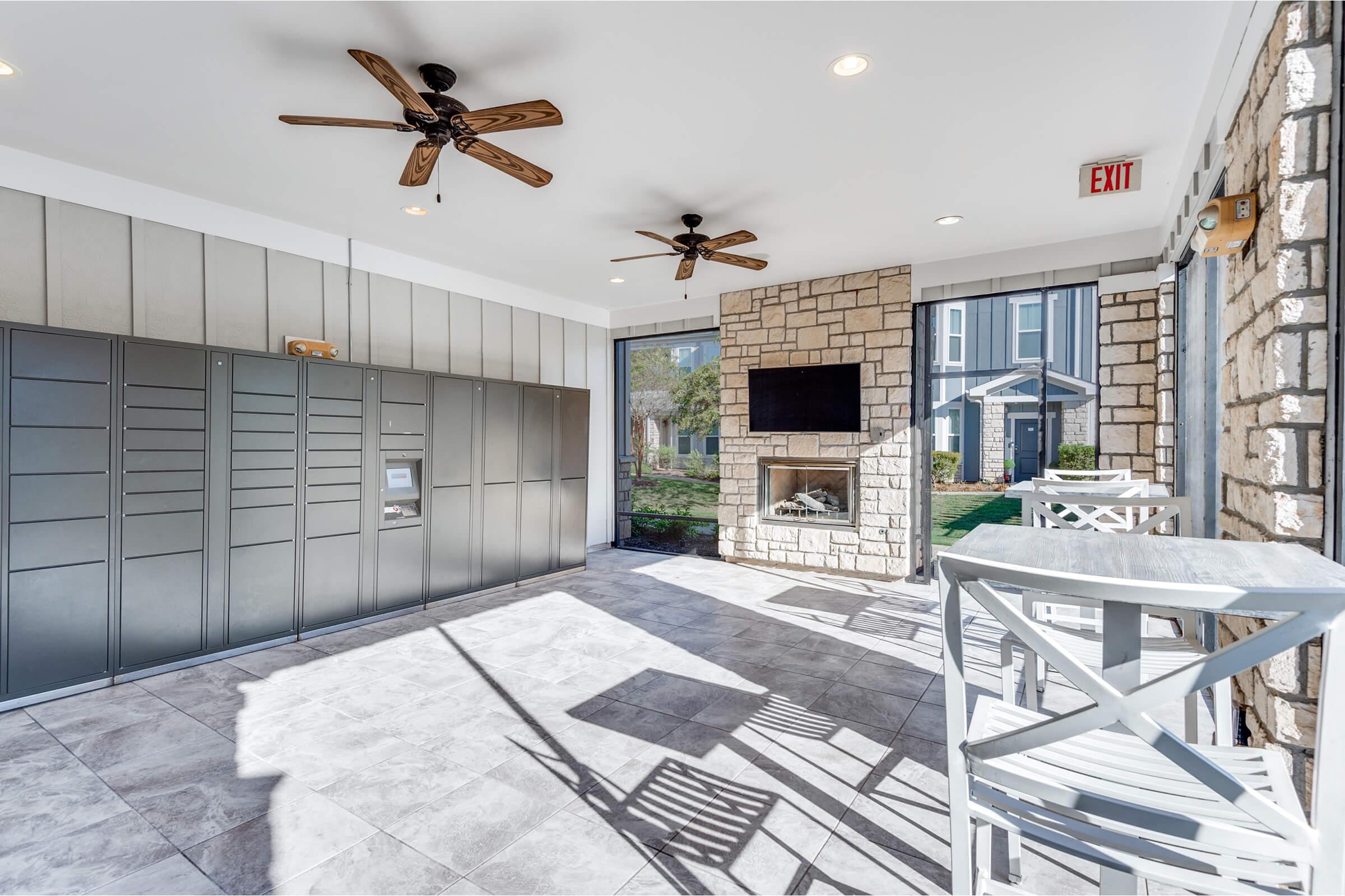 Interior view of a modern recreation room featuring decorative stone walls, two ceiling fans, a television mounted above a fireplace, and a set of white bar-style chairs. Mail lockers are visible along one wall, and large windows allow natural light and a view of the outside.