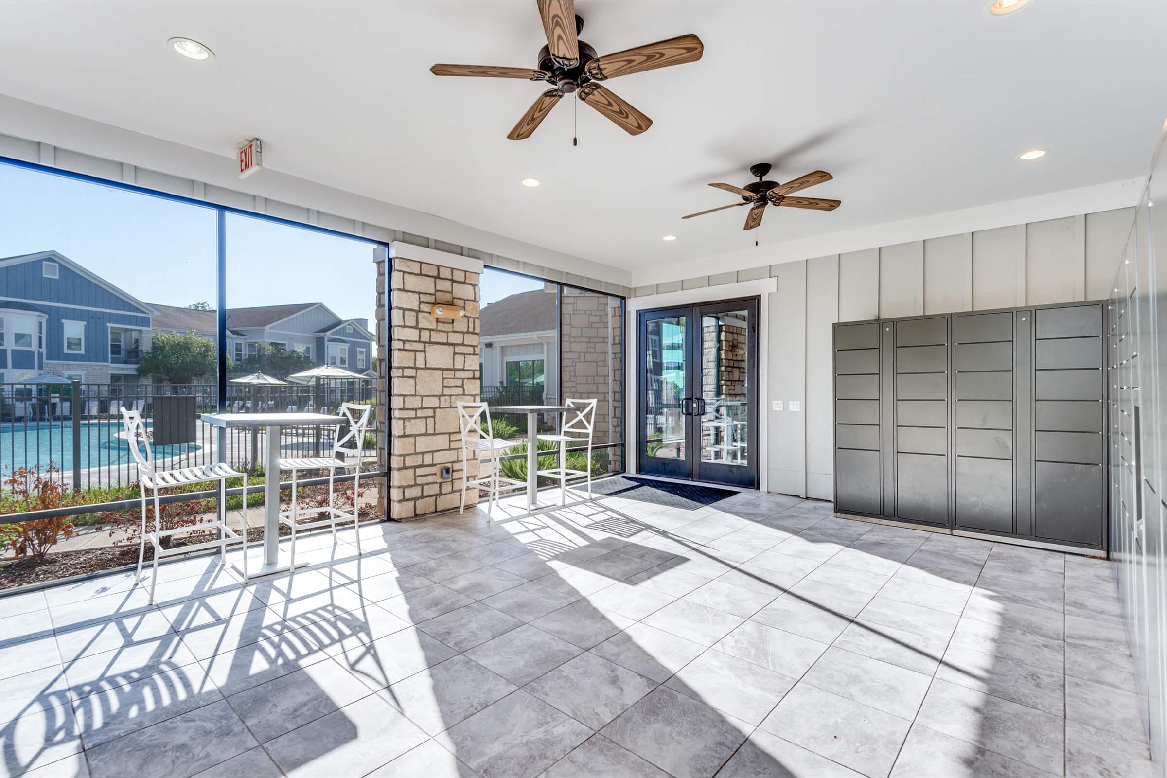 Bright and airy indoor space featuring large windows, stylish ceiling fans, and modern tiled flooring. The area includes seating with high tables and overlooks a pool area outside, creating a welcoming atmosphere. The combination of natural light and contemporary design enhances the overall ambiance.