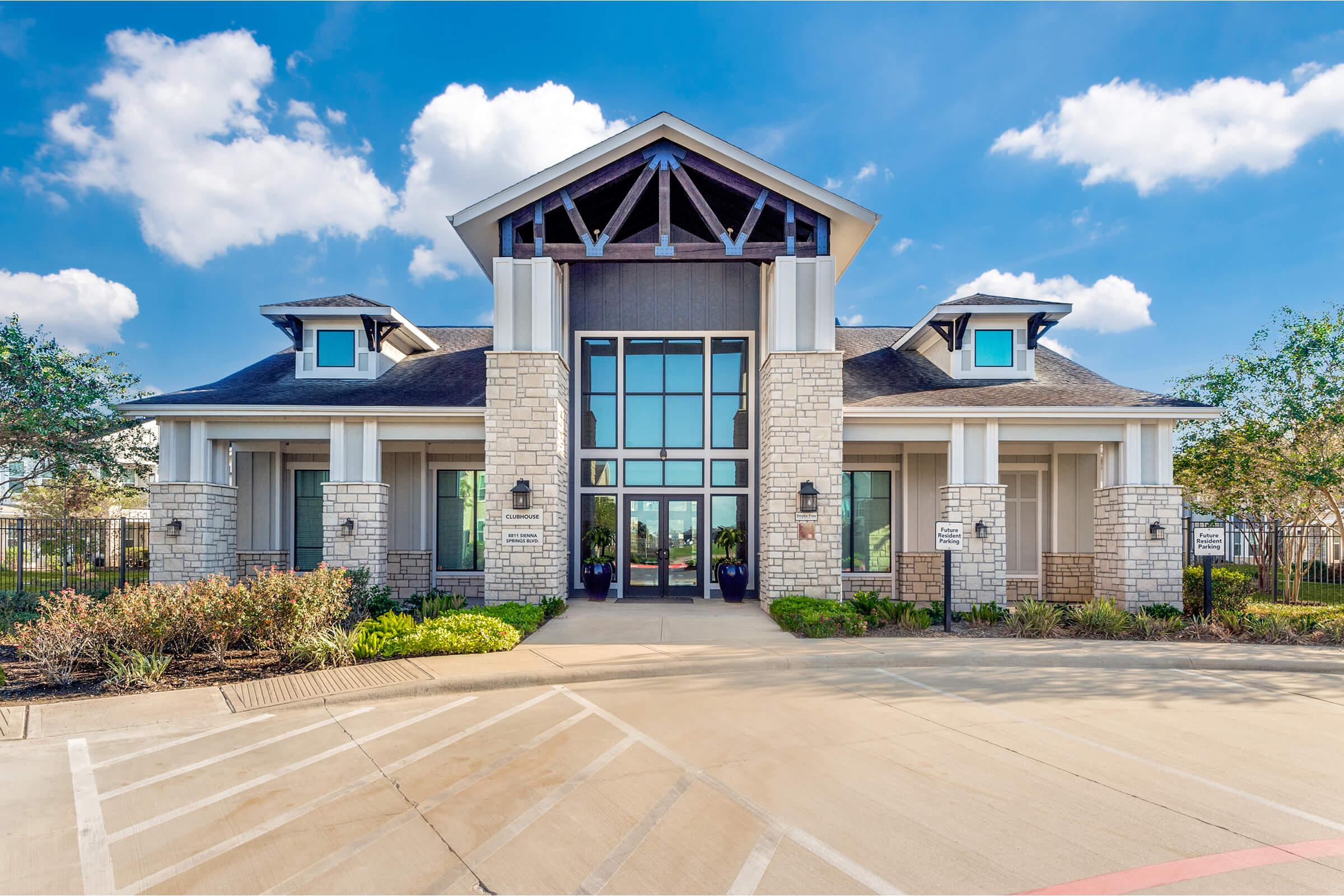 A modern building with a stone and siding exterior, featuring large glass windows and a prominent entrance. The structure has a peaked roof and is surrounded by landscaped greenery. The sky is bright with a few clouds, and there is a paved driveway in front.