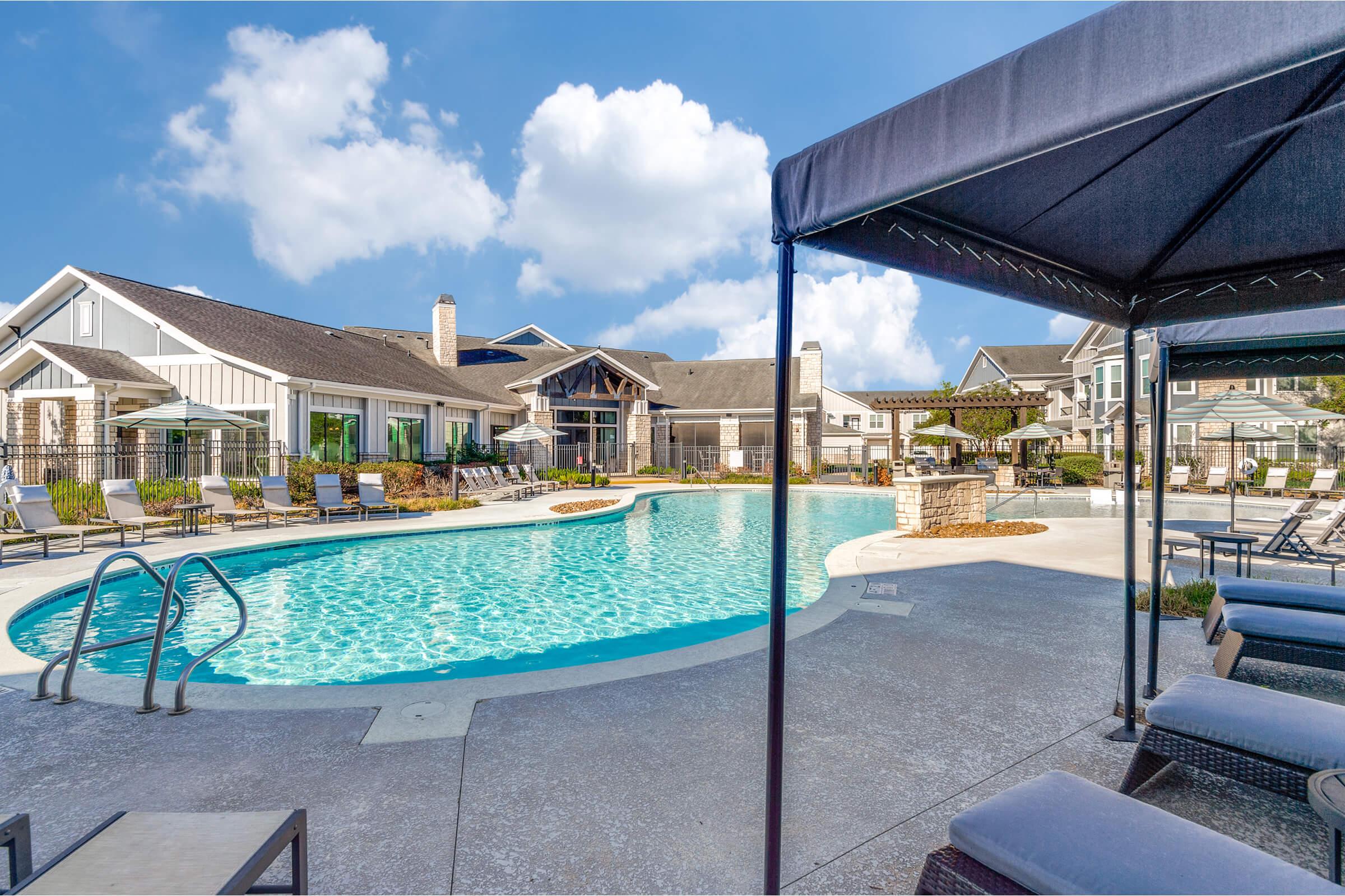 A serene outdoor swimming pool surrounded by lounge chairs and cabanas, featuring a vibrant blue water. In the background, modern residential buildings and lush landscaping under a partly cloudy sky, creating a relaxing atmosphere for leisure and enjoyment.