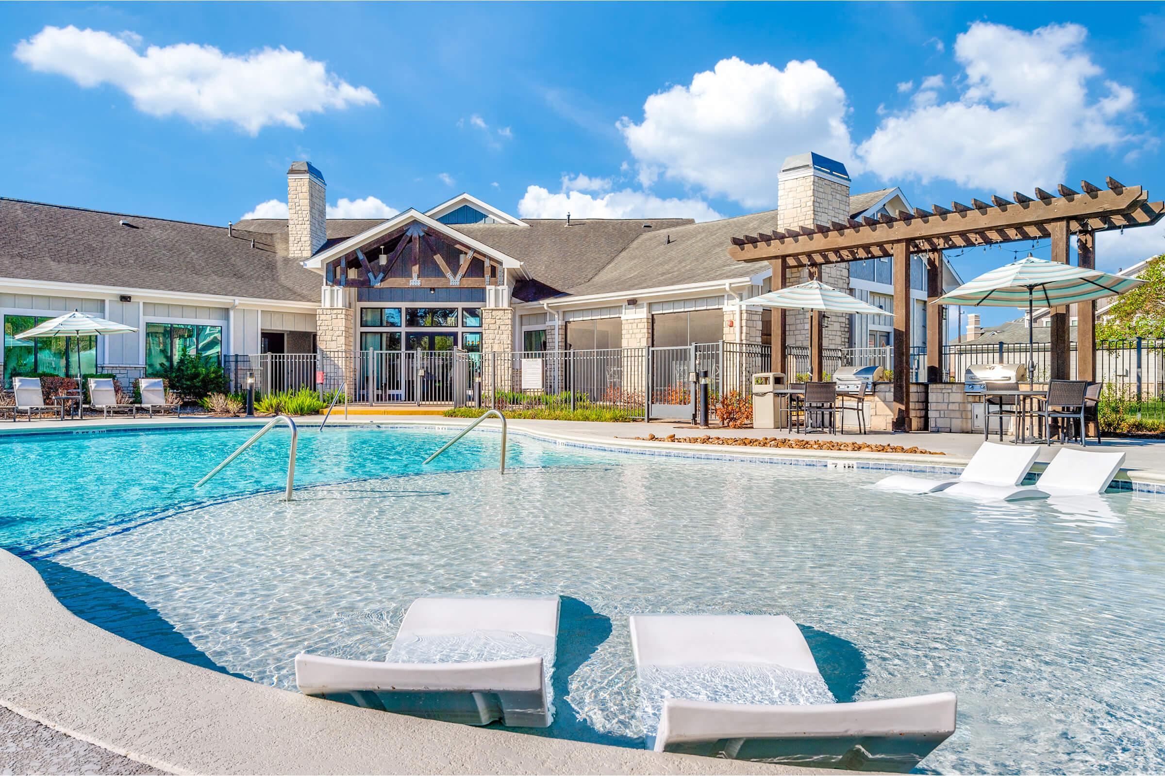 A beautiful outdoor pool features lounge chairs in the foreground, surrounded by a spacious deck. In the background, a modern building with a sloped roof and large windows can be seen, along with shaded seating areas and umbrellas. The sky is bright with fluffy white clouds, creating a relaxing atmosphere.