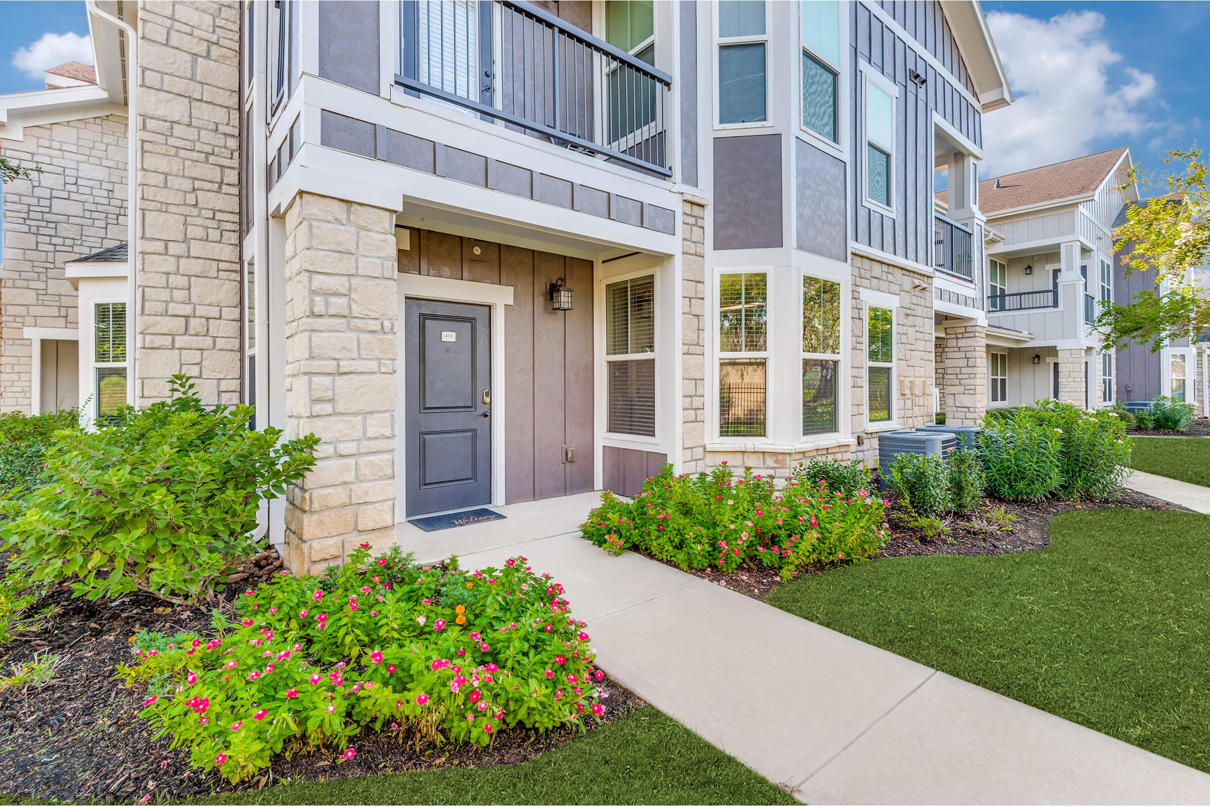 A modern apartment building featuring a stone and siding exterior. The entrance has a dark door with a small light above it, surrounded by shrubs and colorful flowers. A well-maintained path leads to the door, with lush green grass on both sides and a clear blue sky in the background.