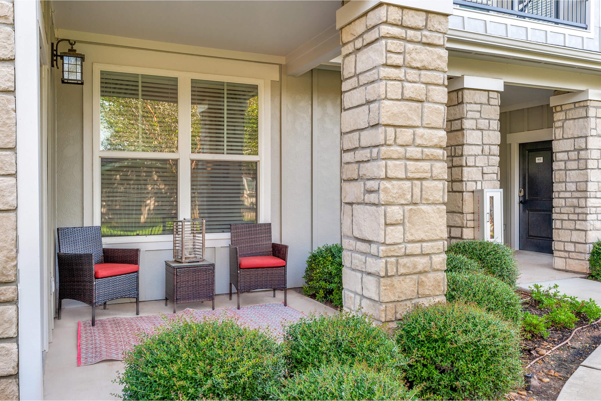 A cozy porch area featuring two woven chairs with red cushions, a small side table, and a decorative lantern. The porch is framed by stone pillars and has lush greenery and manicured bushes around the entrance, creating a welcoming atmosphere.