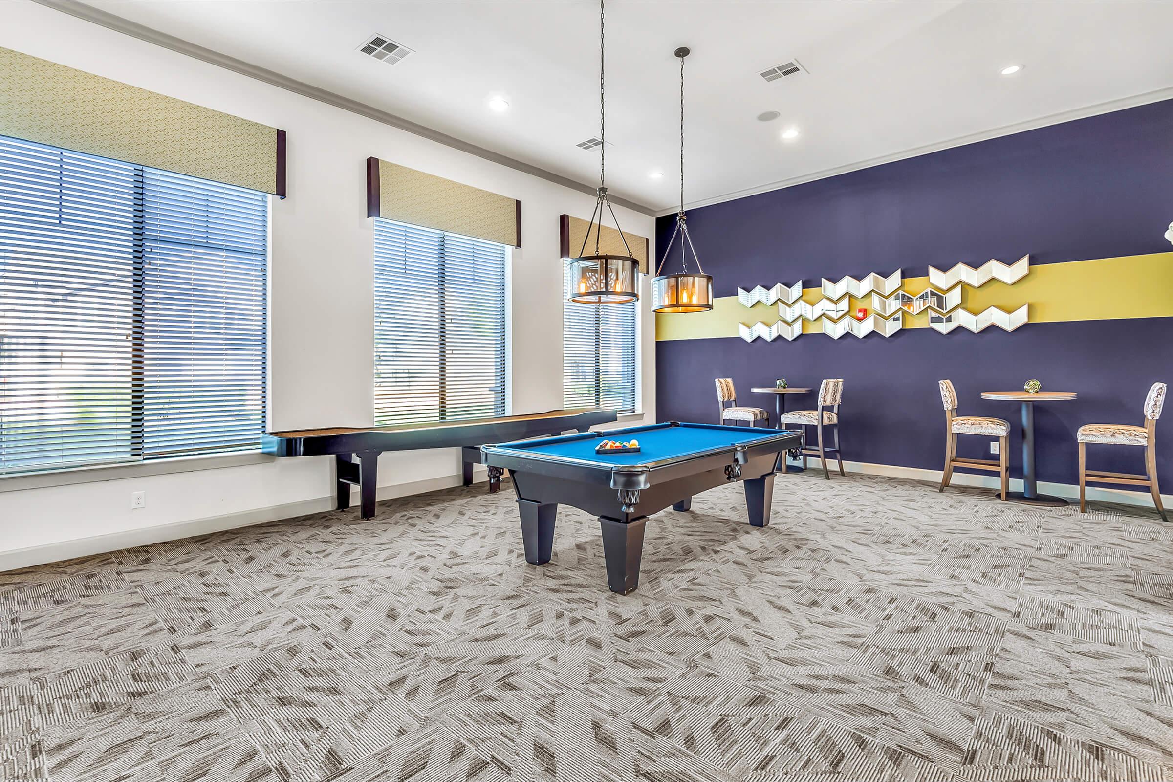 A modern recreational room featuring a blue pool table in the center, flanked by large windows with shades. Two small tables with chairs are visible against the walls, and decorative elements are arranged on one wall, contributing to a stylish and inviting atmosphere.
