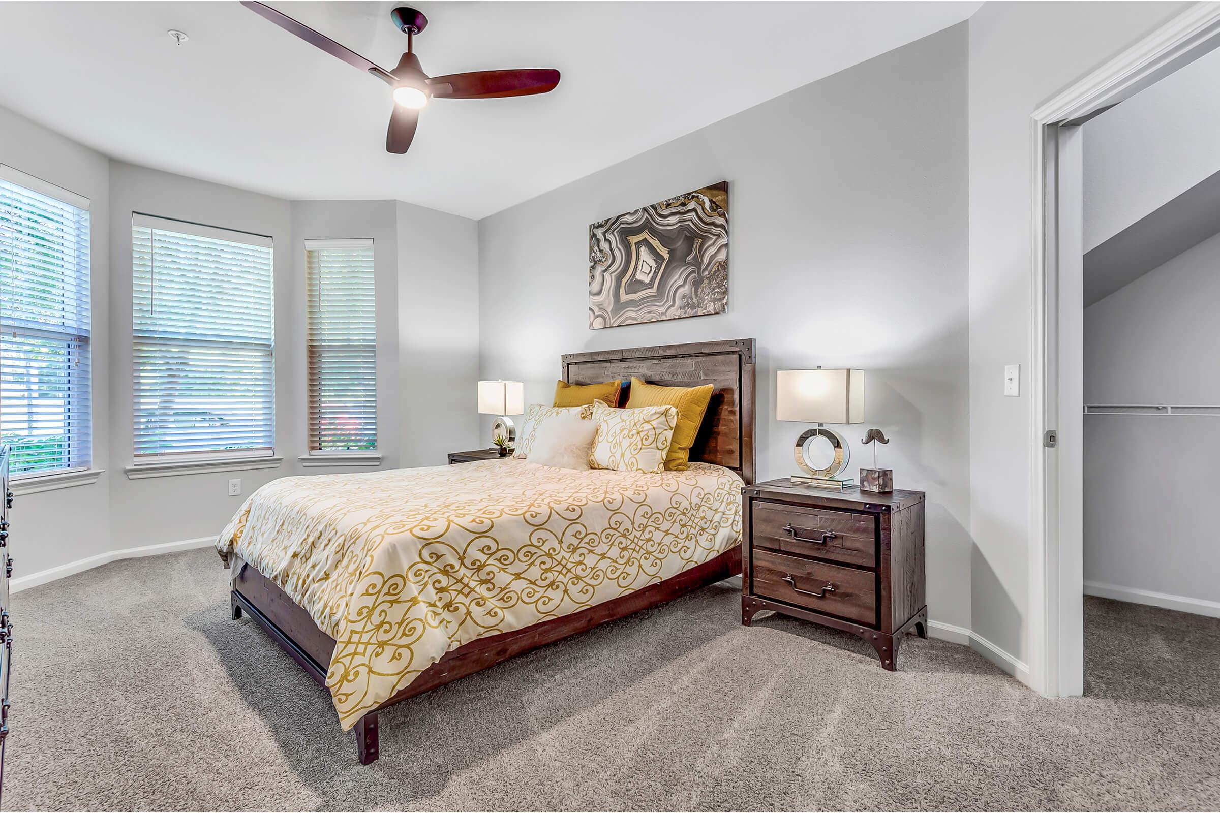 A cozy bedroom featuring a bed with a decorative yellow and gold comforter, two bedside tables with lamps, and a piece of wall art. There are large windows with blinds allowing natural light, and carpeted flooring adds warmth to the space. A door leads to a closet.