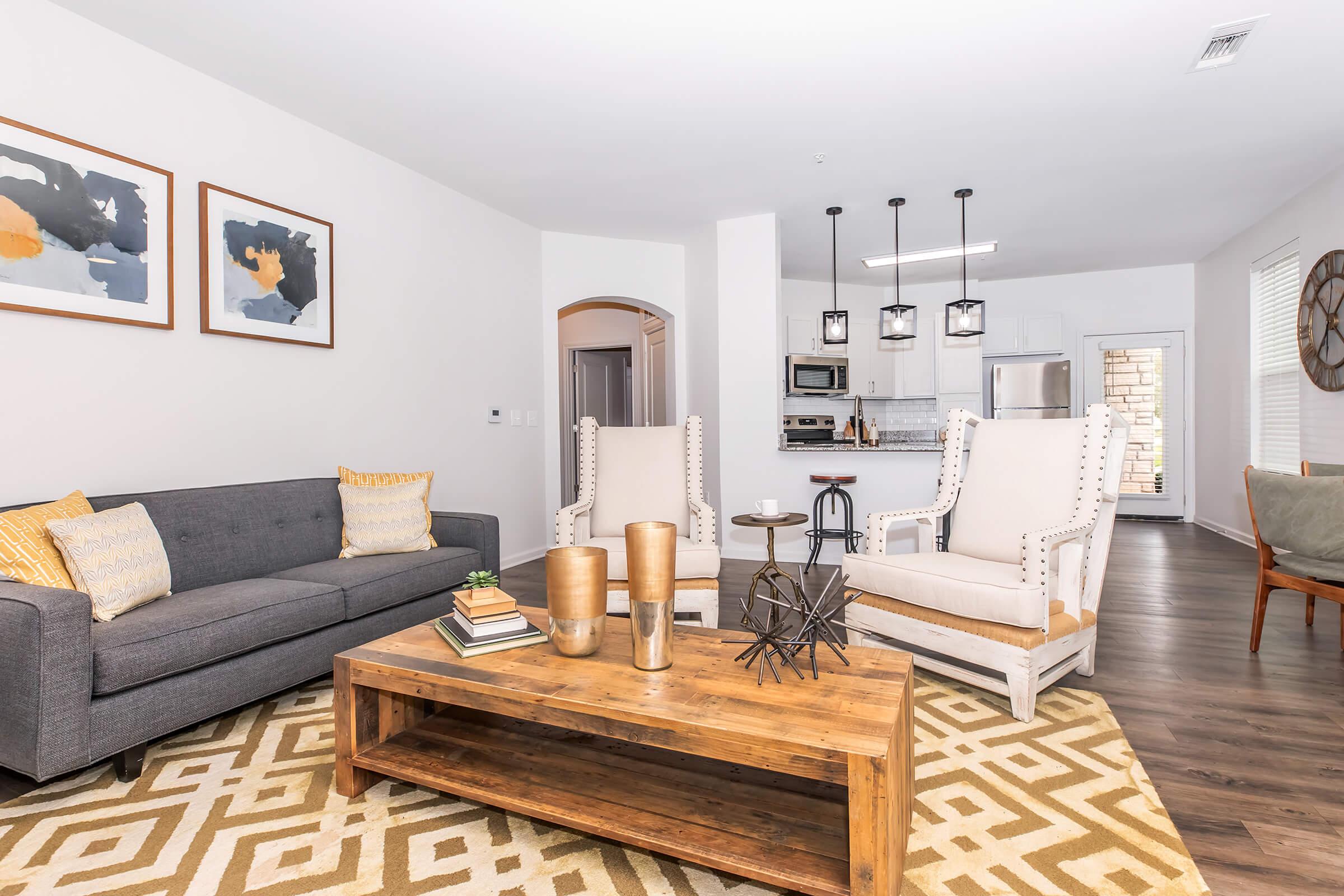 Modern living room featuring a gray sofa with yellow accent pillows, two stylish armchairs, and a wooden coffee table. Decorative elements include books, a plant, and a geometric sculpture. The space has light-colored walls and a cozy atmosphere, with a view of a kitchen in the background.