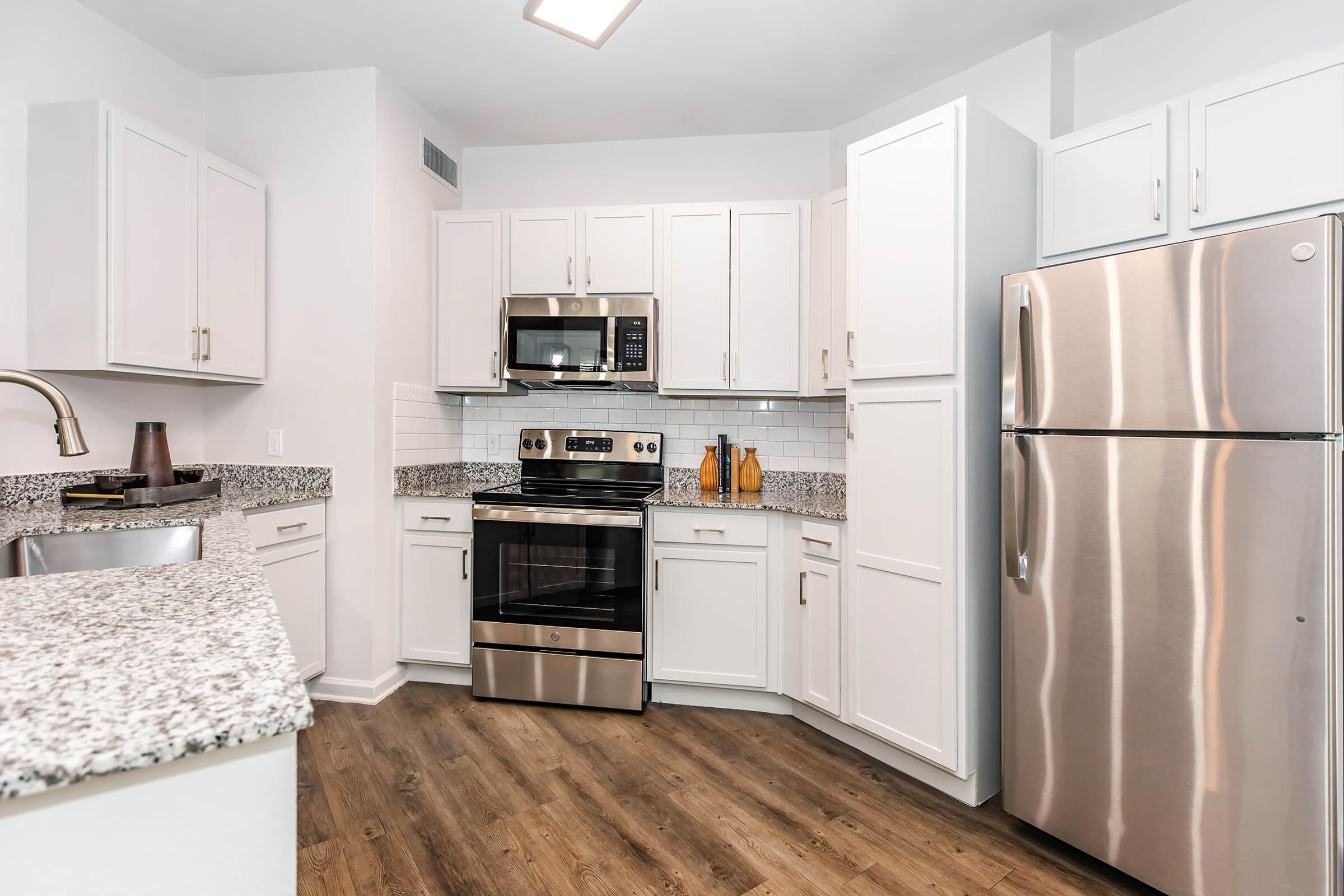 Modern kitchen featuring white cabinetry, stainless steel appliances including a microwave, oven, and refrigerator, complemented by a granite countertop. The space has a light and airy feel with a neutral color palette and wood-style flooring, providing a contemporary culinary environment.