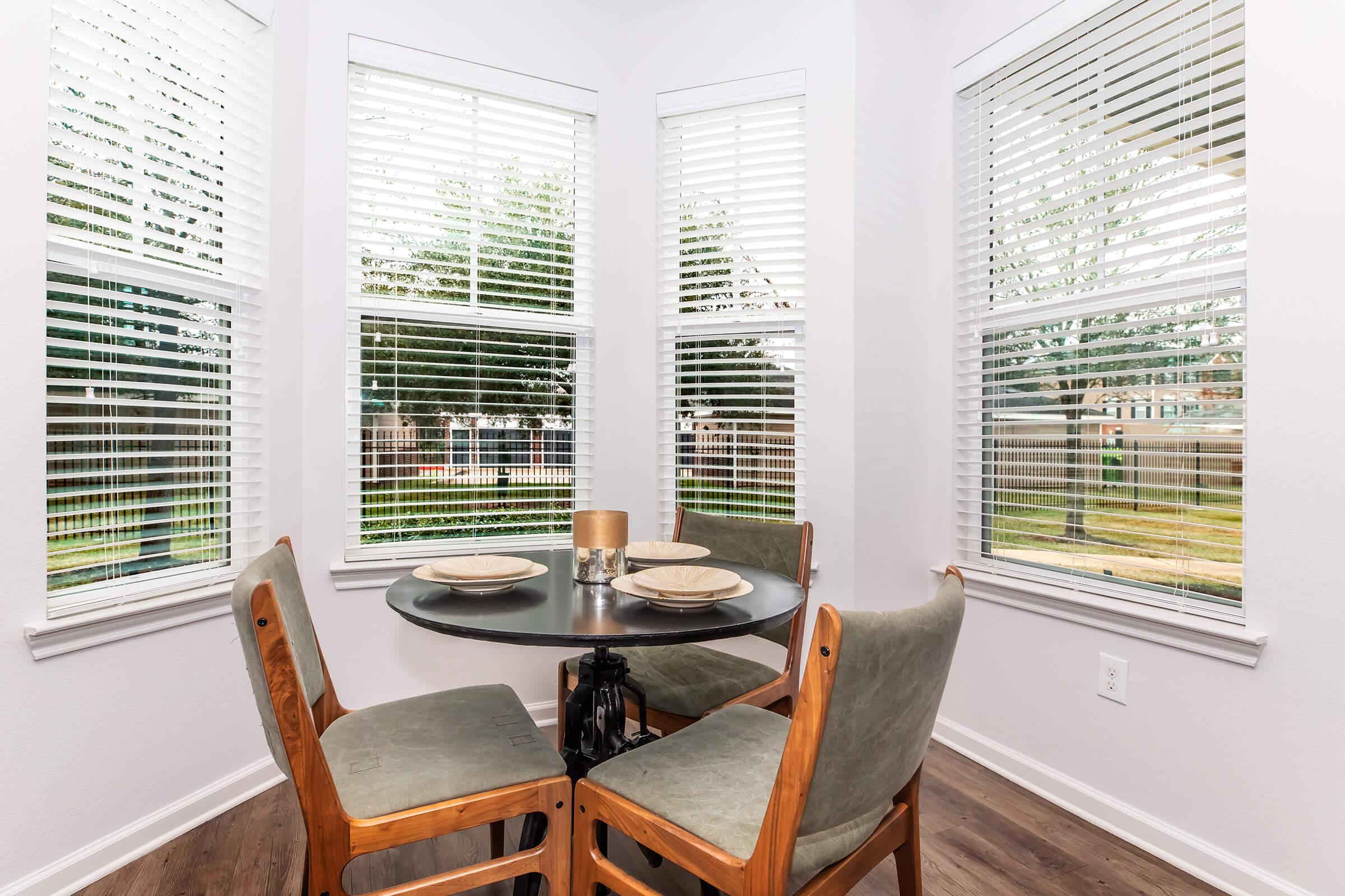 A cozy dining nook featuring a round black table surrounded by four wooden chairs with green cushions. The space is brightened by natural light streaming through large windows adorned with white blinds, offering a view of trees outside. The neutral-colored walls create a calm atmosphere.