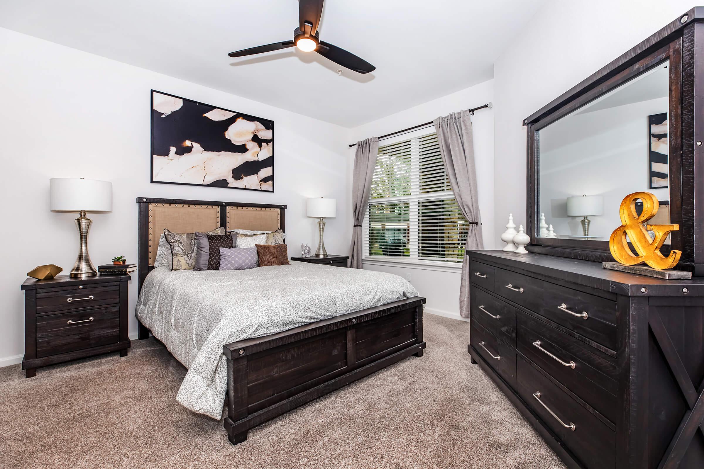 A cozy bedroom featuring a gray upholstered bed with decorative pillows, bedside tables with lamps, a dark wood dresser, and a large mirror. Natural light streams through a window adorned with gray curtains, and a piece of abstract art hangs above the bed, adding a modern touch to the decor.