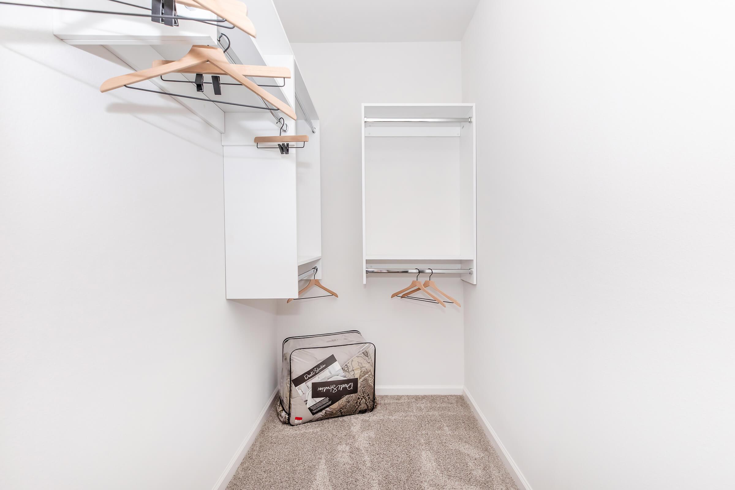 A clean, minimally furnished closet featuring a white wall, a carpeted floor, and two hanging shelves with wooden hangers. A black and white suitcase is positioned on the floor, suggesting a tidy and organized storage space.