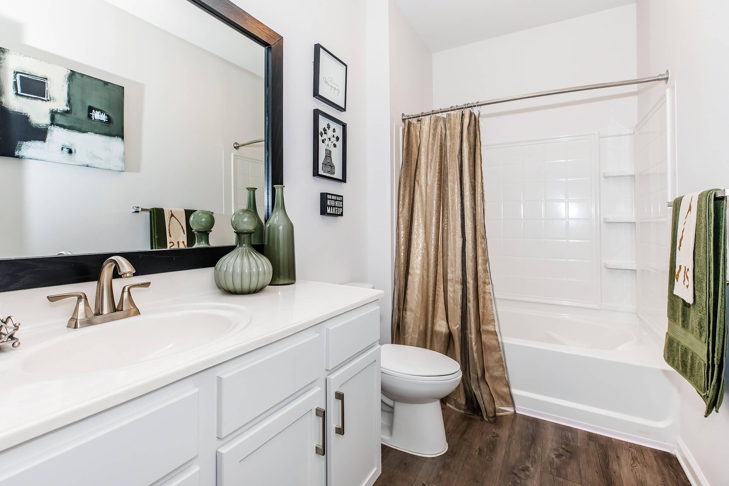 A modern bathroom featuring a white vanity with a sink, a large mirror with a black frame, and decorative items on the counter. There's a bathtub with a beige curtain, a green towel, and art on the wall. The flooring is dark wood, adding a contemporary touch to the space.