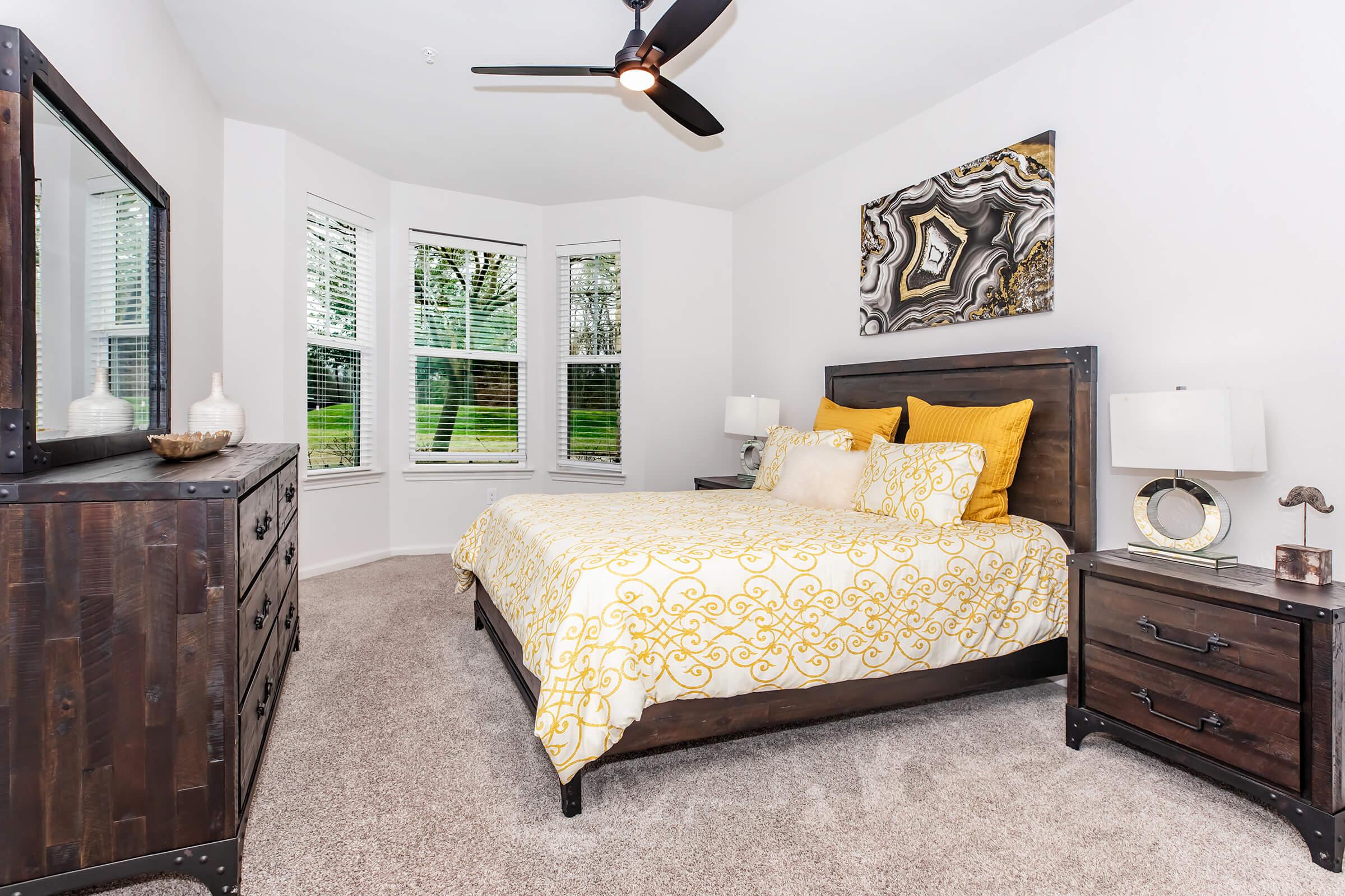 A cozy bedroom featuring a large bed with a yellow and white patterned comforter, two bedside tables with lamps, a dresser, and a large window showcasing green outdoor views. The room has a modern aesthetic with a ceiling fan and decorative artwork on the wall.