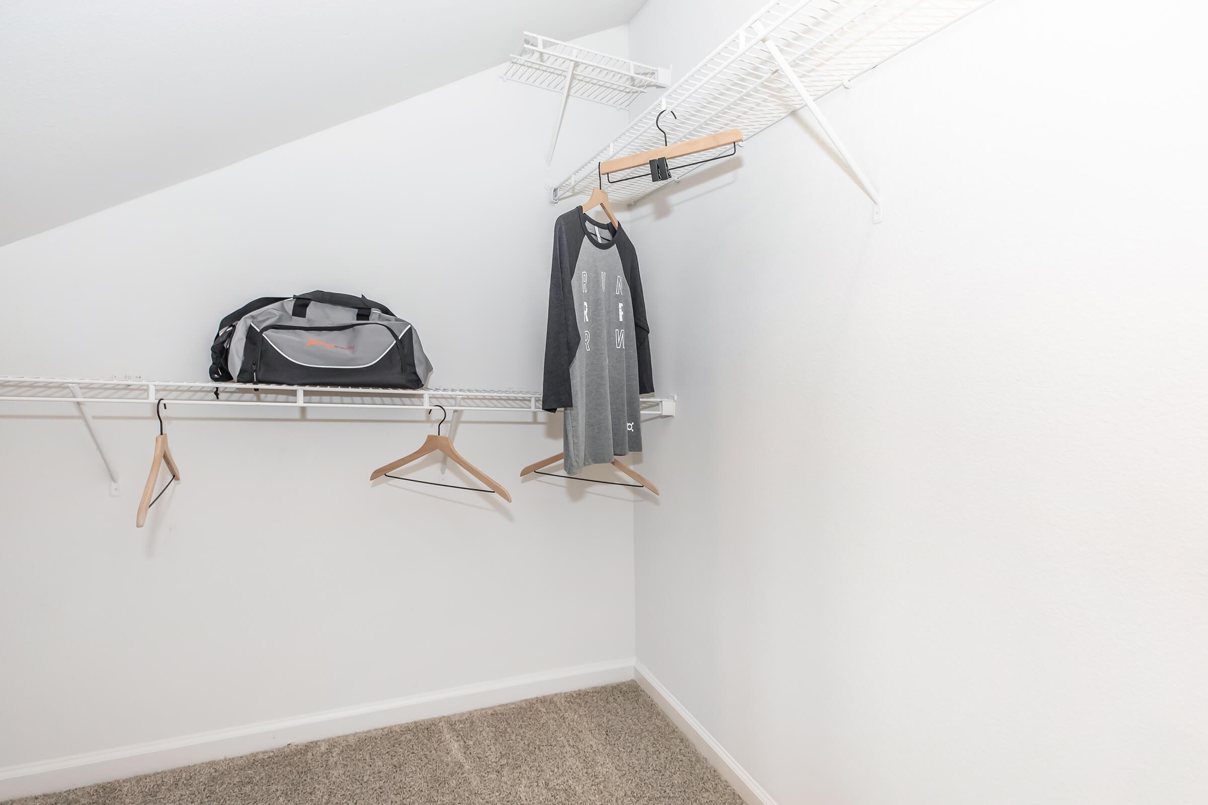 A minimalist closet with white walls and a carpeted floor. It features an open design with two hanging rods, one holding a gray long-sleeve shirt on a hanger and the other with a few empty hangers. A black duffel bag sits on the floor beneath the hanging clothes.