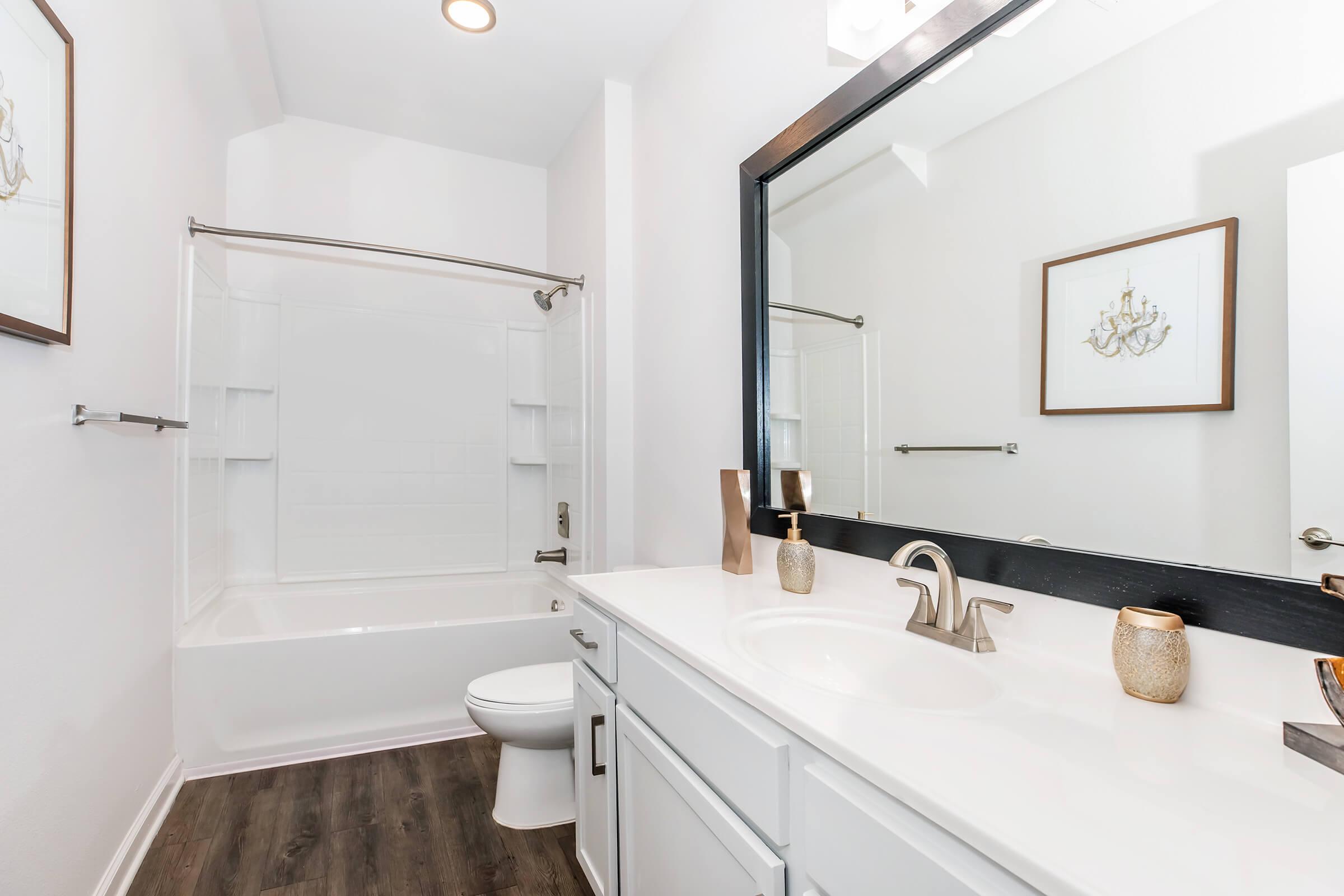 A modern bathroom featuring a white bathtub and shower combination, a sleek vanity with a sink, bathroom accessories, and a large mirror. The walls are painted in a light color, and there is a decorative framed picture on the wall, adding a touch of elegance to the space.