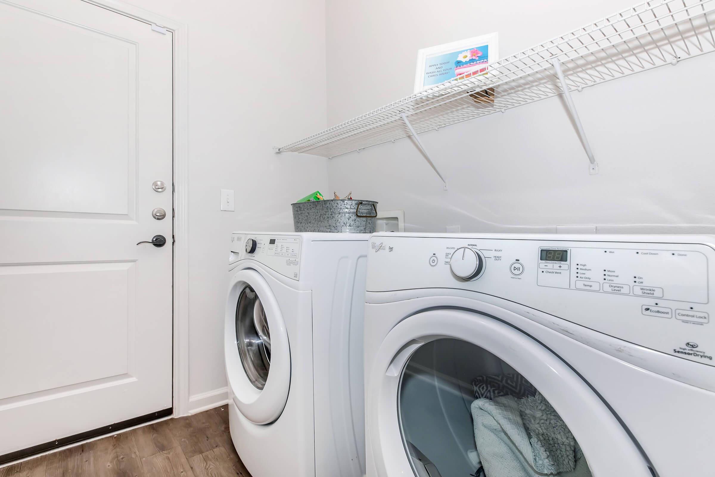 A clean laundry room featuring a set of white front-loading washing machines side by side. Above the machines is a wire shelf holding a metal basket and other items. The room has a light wall color and a door leading outside, with wooden flooring enhancing the spacious appearance.