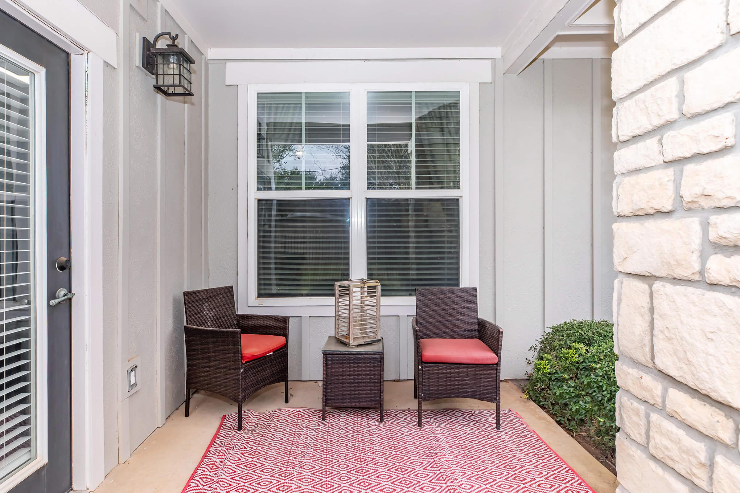 A cozy outdoor seating area featuring two wicker chairs with red cushions, a decorative lantern on a small table, and a patterned red rug. The space is framed by a stone wall and large window with white blinds, surrounded by greenery.