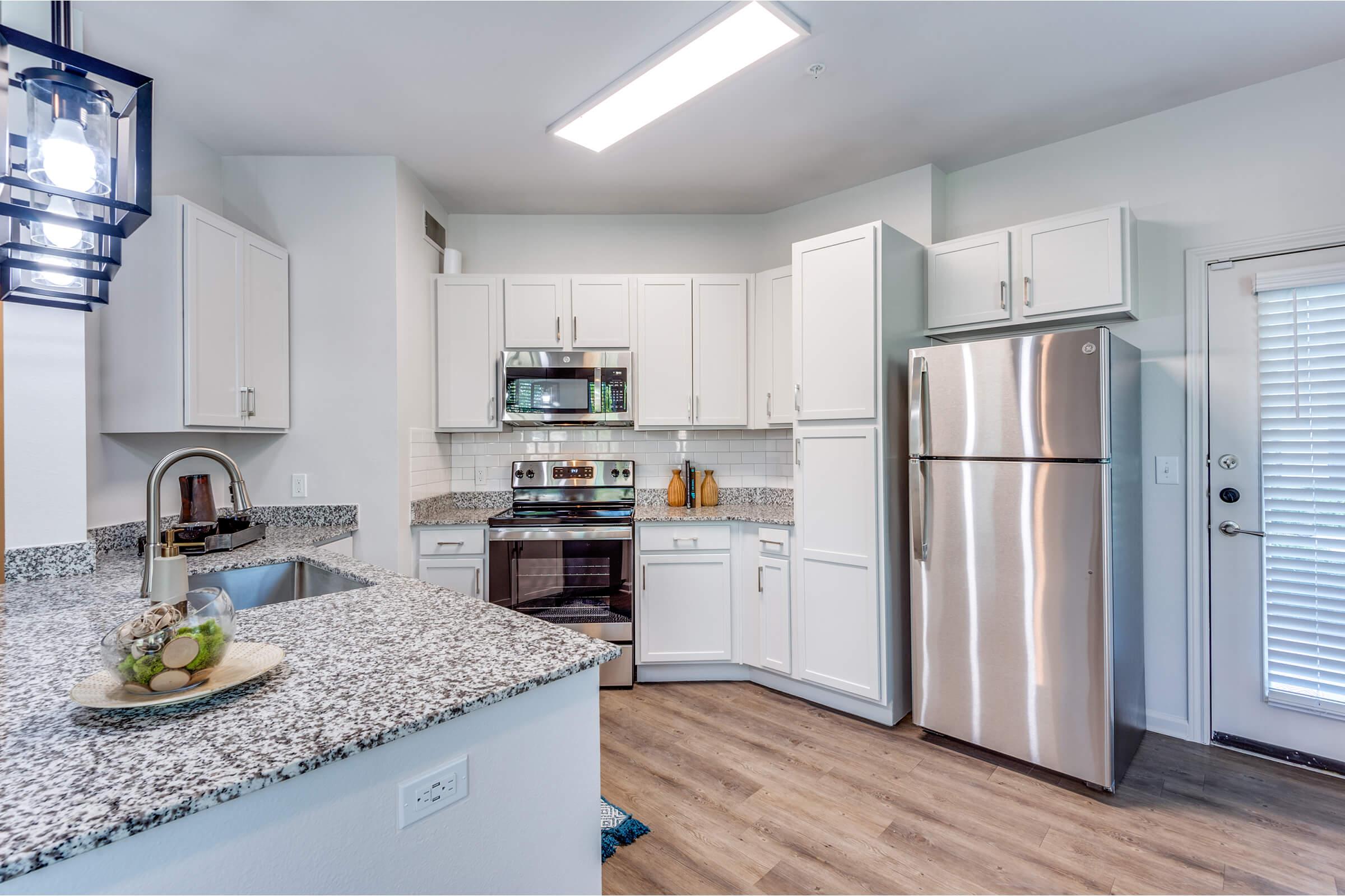 Modern kitchen featuring white cabinetry, stainless steel appliances including a refrigerator and oven, and a granite countertop. The space is well-lit with overhead lighting and has a door leading outside. The flooring is a warm wood finish, complementing the clean, contemporary design.