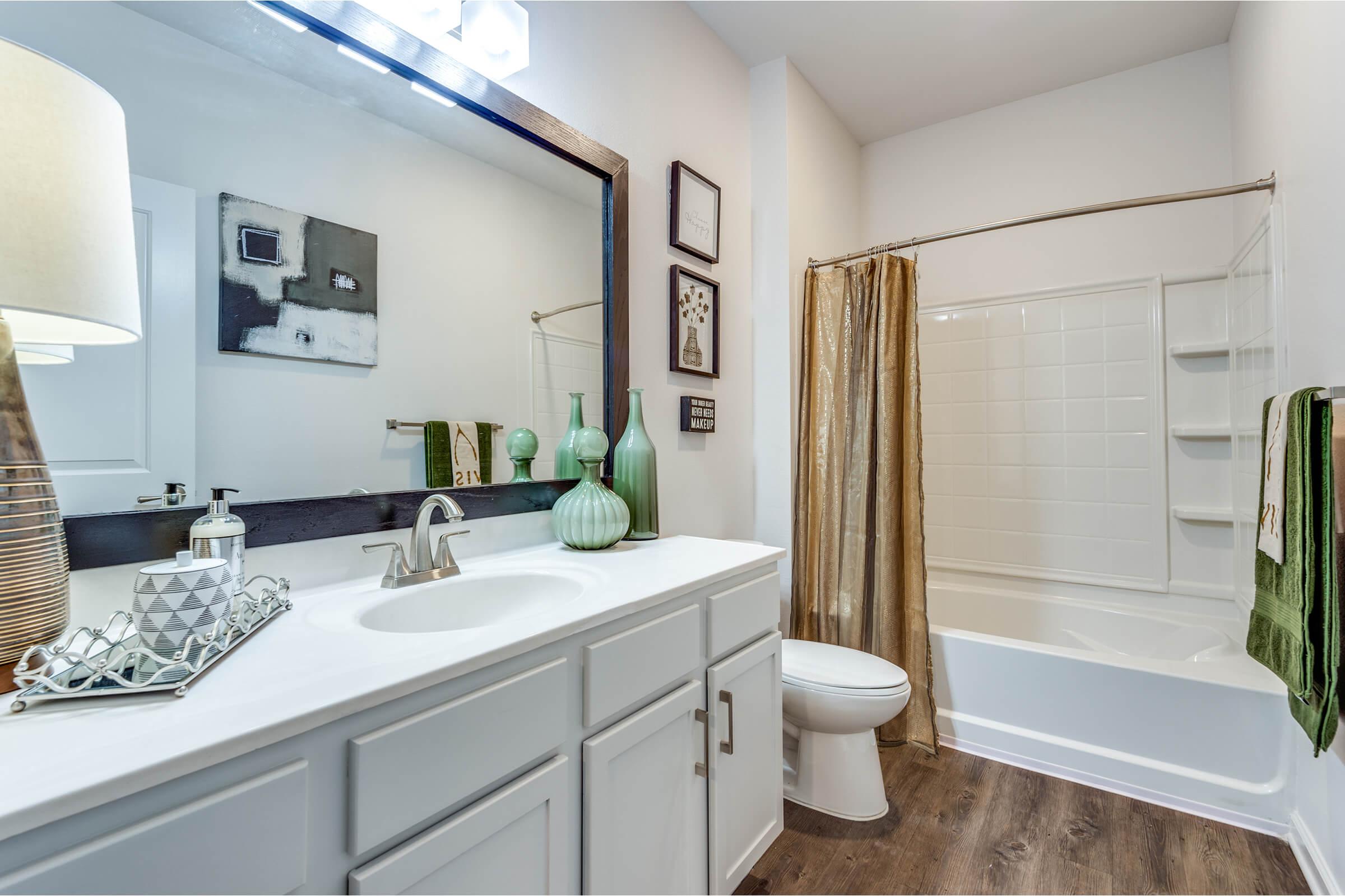 A modern bathroom featuring a spacious vanity with a large mirror, a white bathtub, and a shower area. The decor includes green and gray accents, a lamp, an assortment of decorative bottles, and a geometric tray. Warm wooden flooring complements the bright, neutral walls.