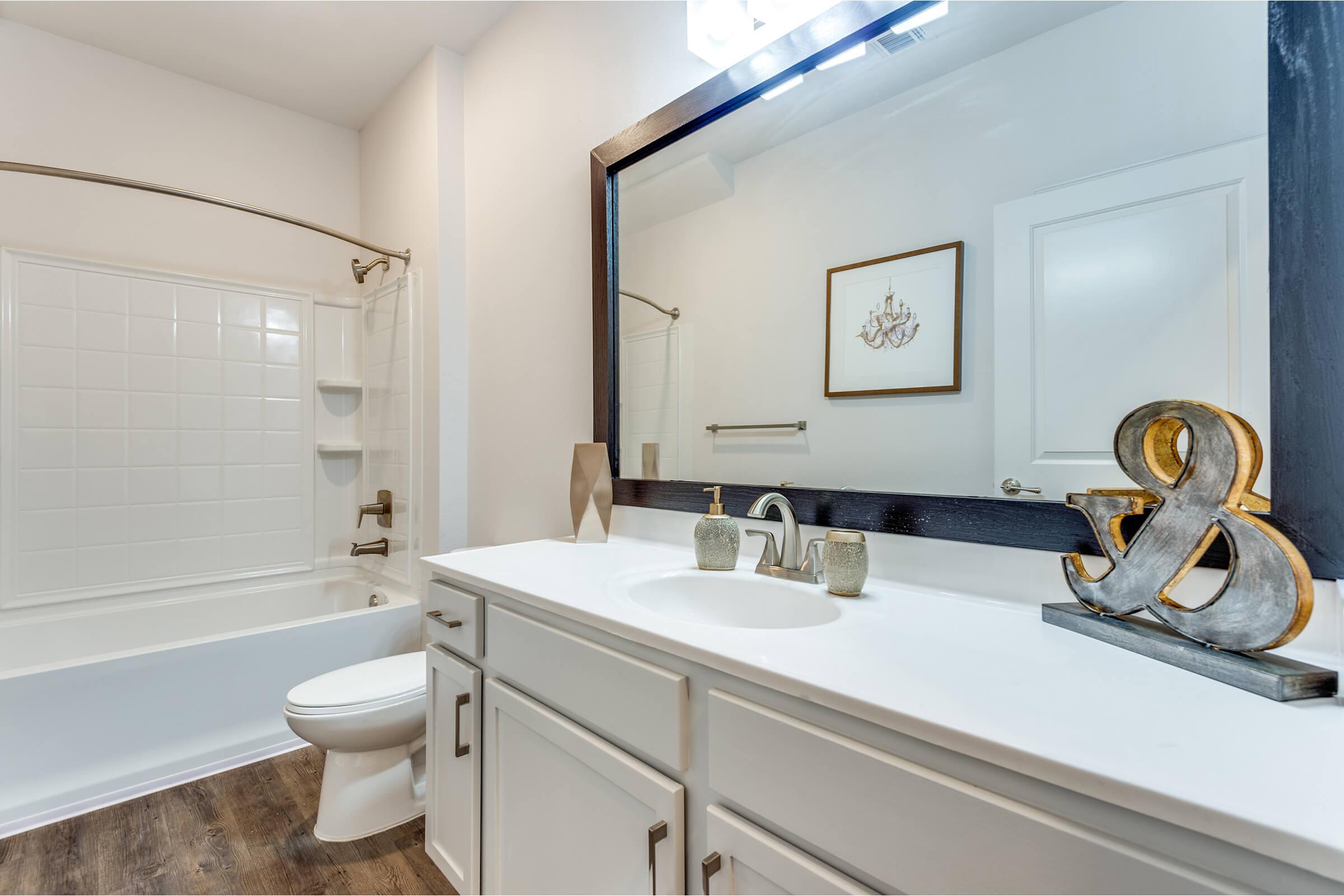 Modern bathroom featuring a white vanity with a sink, decorative items, a large mirror, and a bathtub with an overhead shower. The walls are painted in a light color, and there are stylish accents like a framed chandelier and an ampersand sculpture on the counter. Wood-like flooring adds warmth to the space.