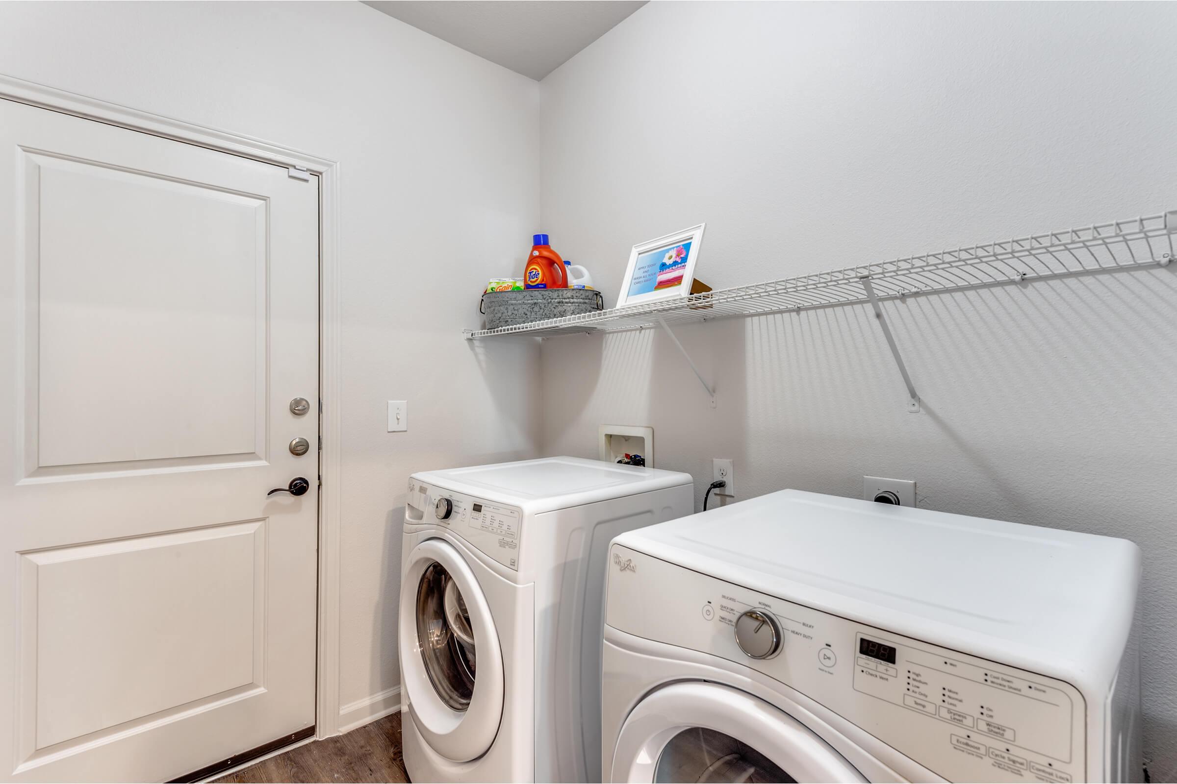 A bright laundry room featuring a white washer and dryer side by side. There's a door on the left leading outside and a wall-mounted shelf above with a basket and a framed picture. The walls are painted in a light shade, creating an airy and clean atmosphere.