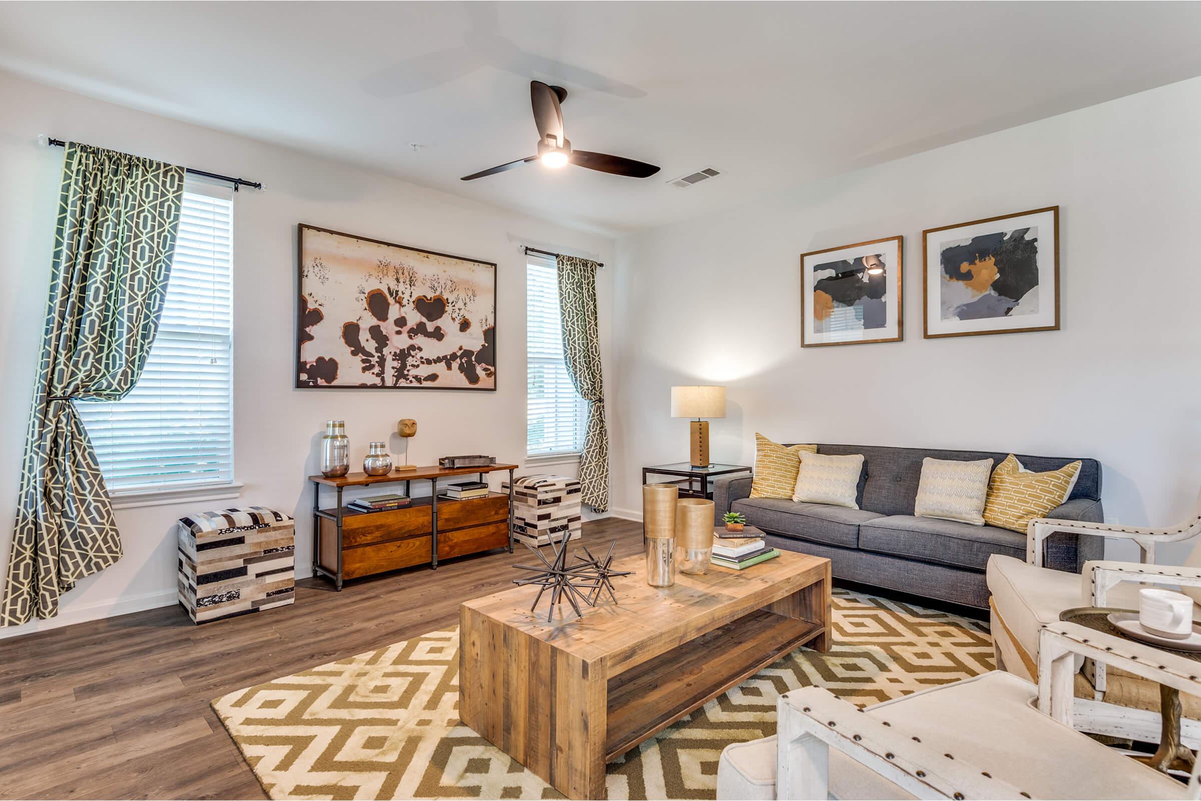 A stylish living room featuring a gray sofa with decorative pillows, a wooden coffee table, and an accent table with a lamp. Walls are adorned with abstract artwork. Floor-to-ceiling curtains frame two windows, and a patterned area rug adds warmth to the space. Storage baskets are placed nearby for decor.