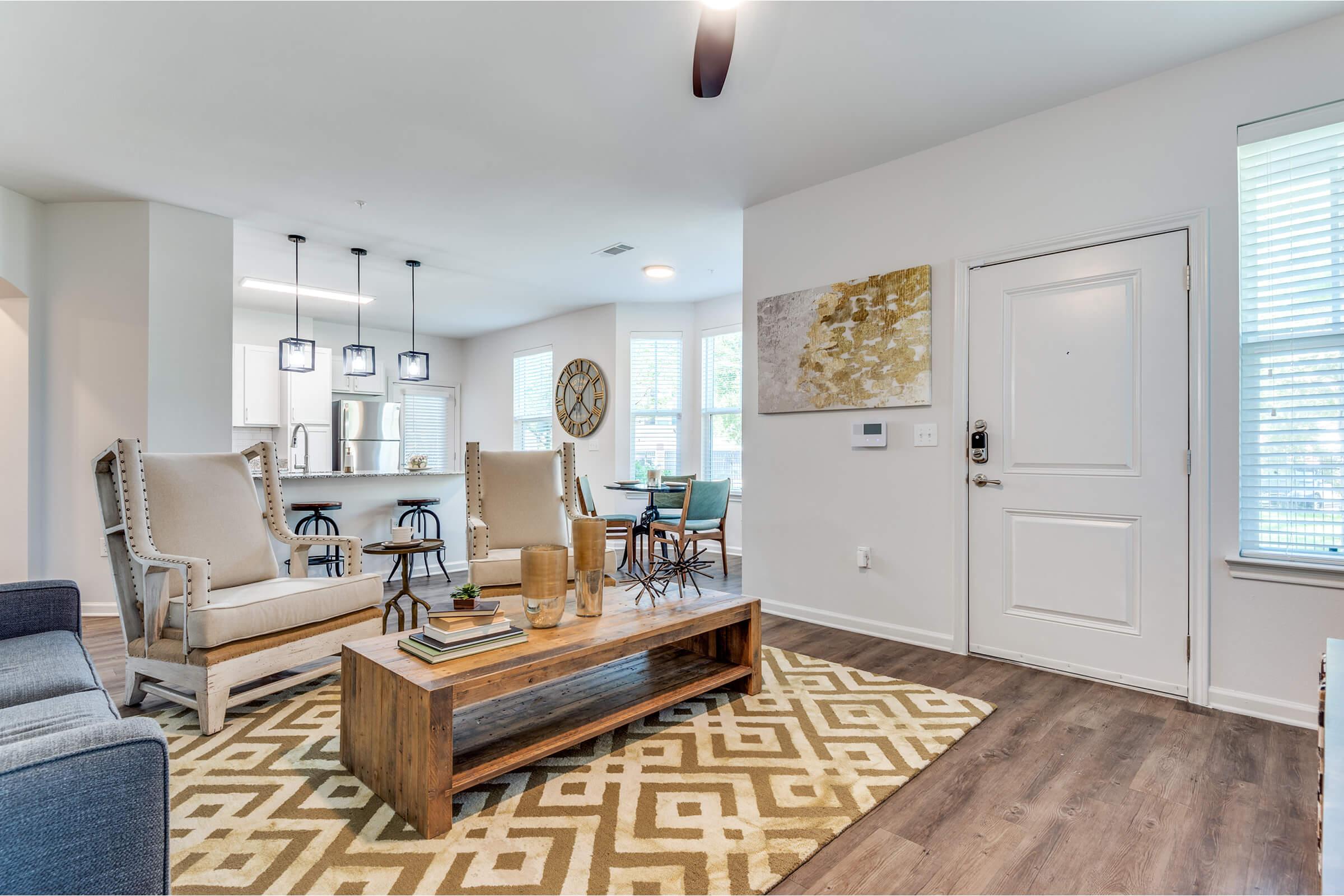 A modern living room featuring two stylish armchairs, a wooden coffee table on a geometric area rug, and large windows allowing natural light. A dining area is visible in the background with pendant lighting and a round clock. The decor is minimal and contemporary, creating a welcoming atmosphere.