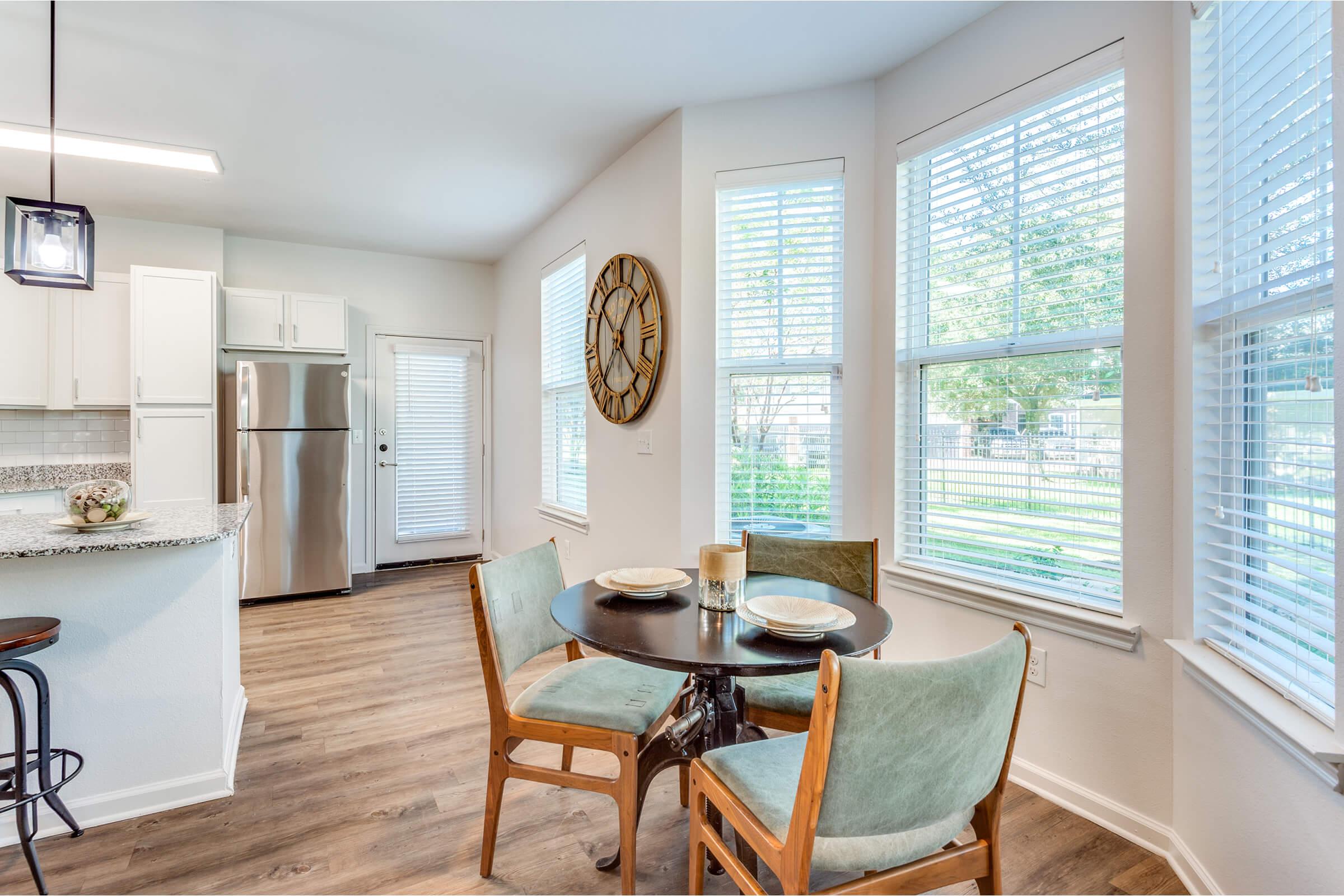 A bright kitchen area featuring a small round dining table with two plates and glasses. Surrounding the table are wooden chairs with green upholstery. The kitchen has modern appliances, including a silver refrigerator, and large windows allowing natural light to fill the space. A wooden wall clock adds a decorative touch.