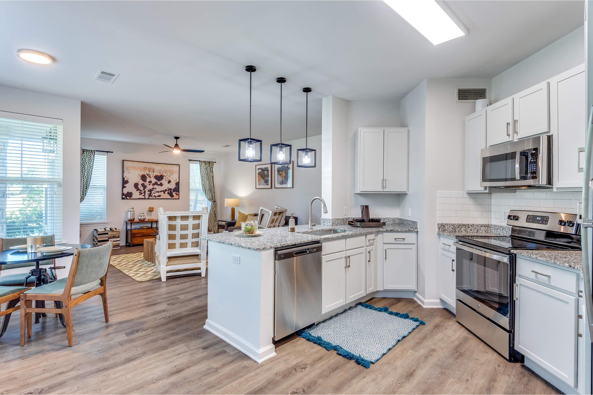A modern kitchen featuring white cabinets, stainless steel appliances, and a granite countertop. A dining area with a round table and chairs is visible, along with a cozy living room space in the background, adorned with a rug and stylish decor. Natural light pours in through the windows.