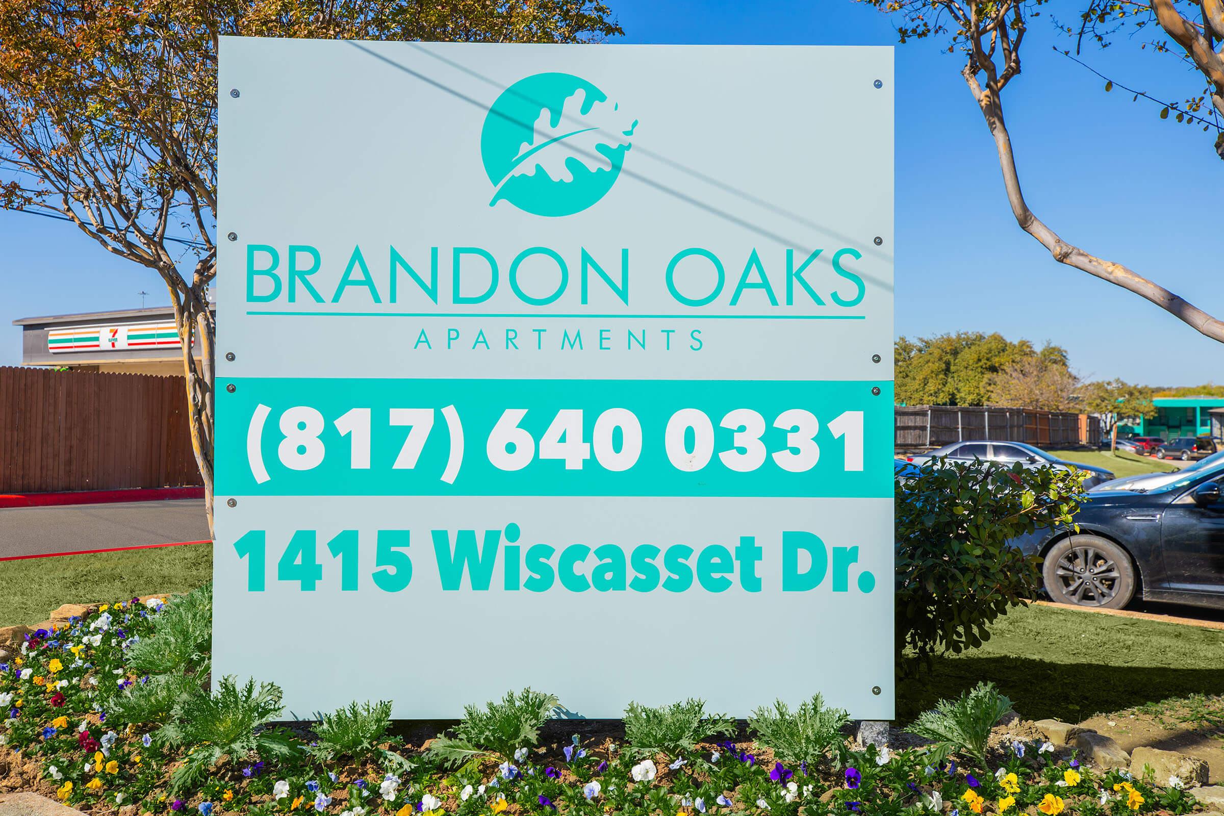 Sign for Brandon Oaks Apartments displaying contact information: phone number (817) 640-0331 and address 1415 Wiscasset Dr. Surrounded by decorative landscaping with a clear blue sky in the background.