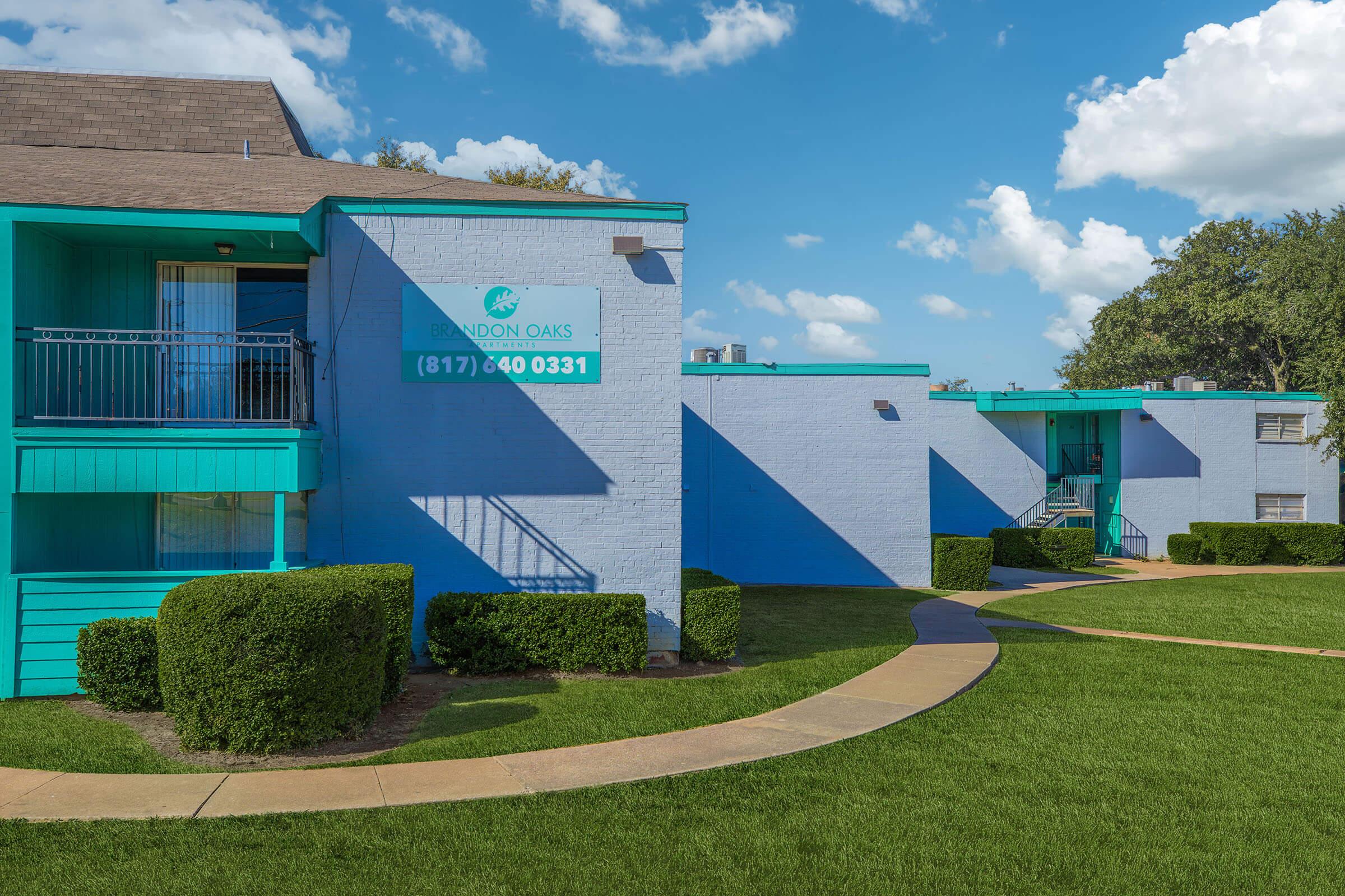 Exterior view of a light blue apartment building with teal accents. The building features a sign displaying the name "Brandon Oaks" with a phone number. Neatly trimmed shrubs line the walkway leading up to the building. There are white clouds in a blue sky, creating a bright and inviting atmosphere.