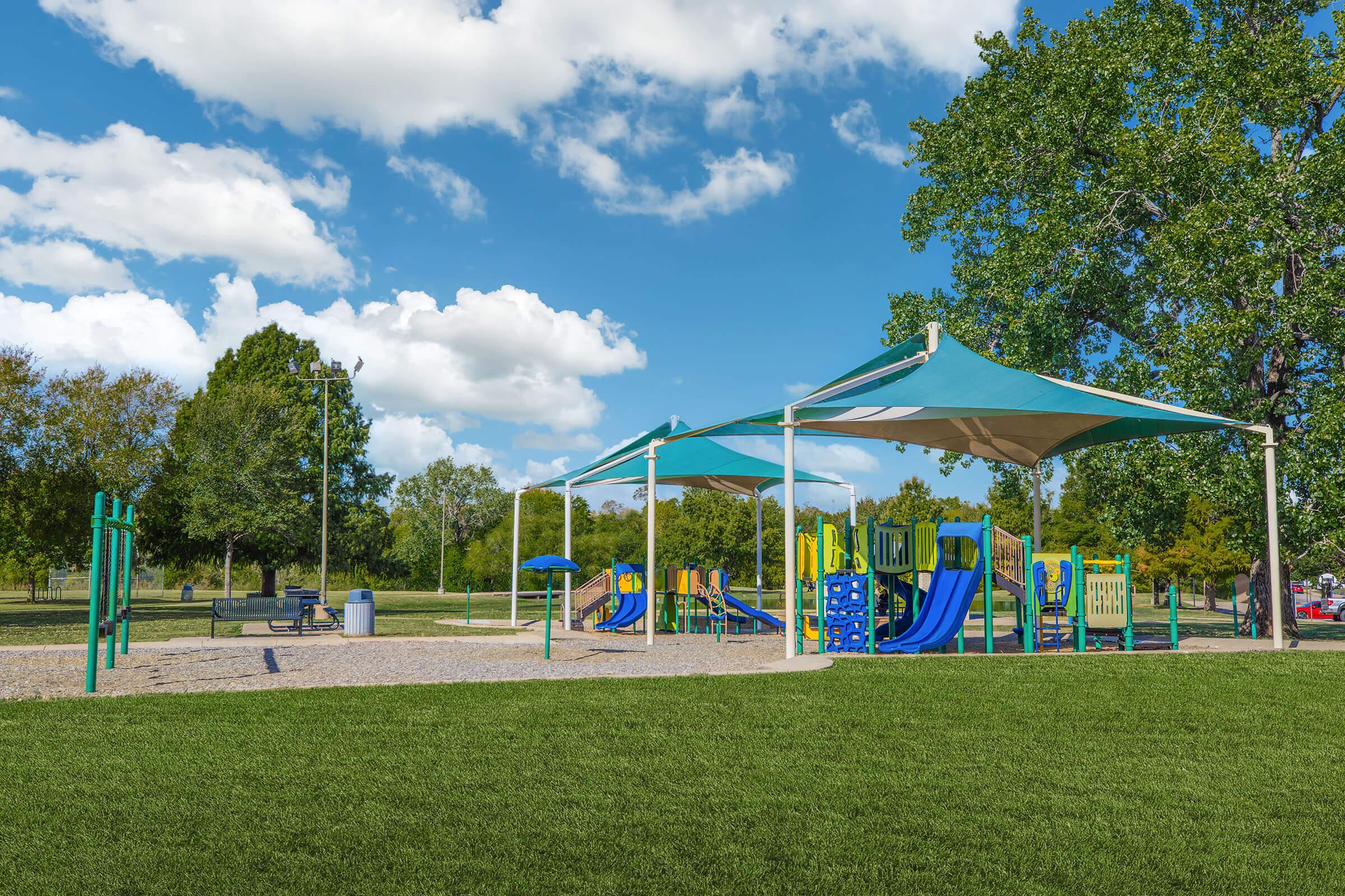 Playground scene featuring colorful play structures, slides, and climbing areas under a large shade canopy. Surrounded by green grass, trees, and a bright blue sky with fluffy clouds, the location offers a cheerful environment for children to play and explore.