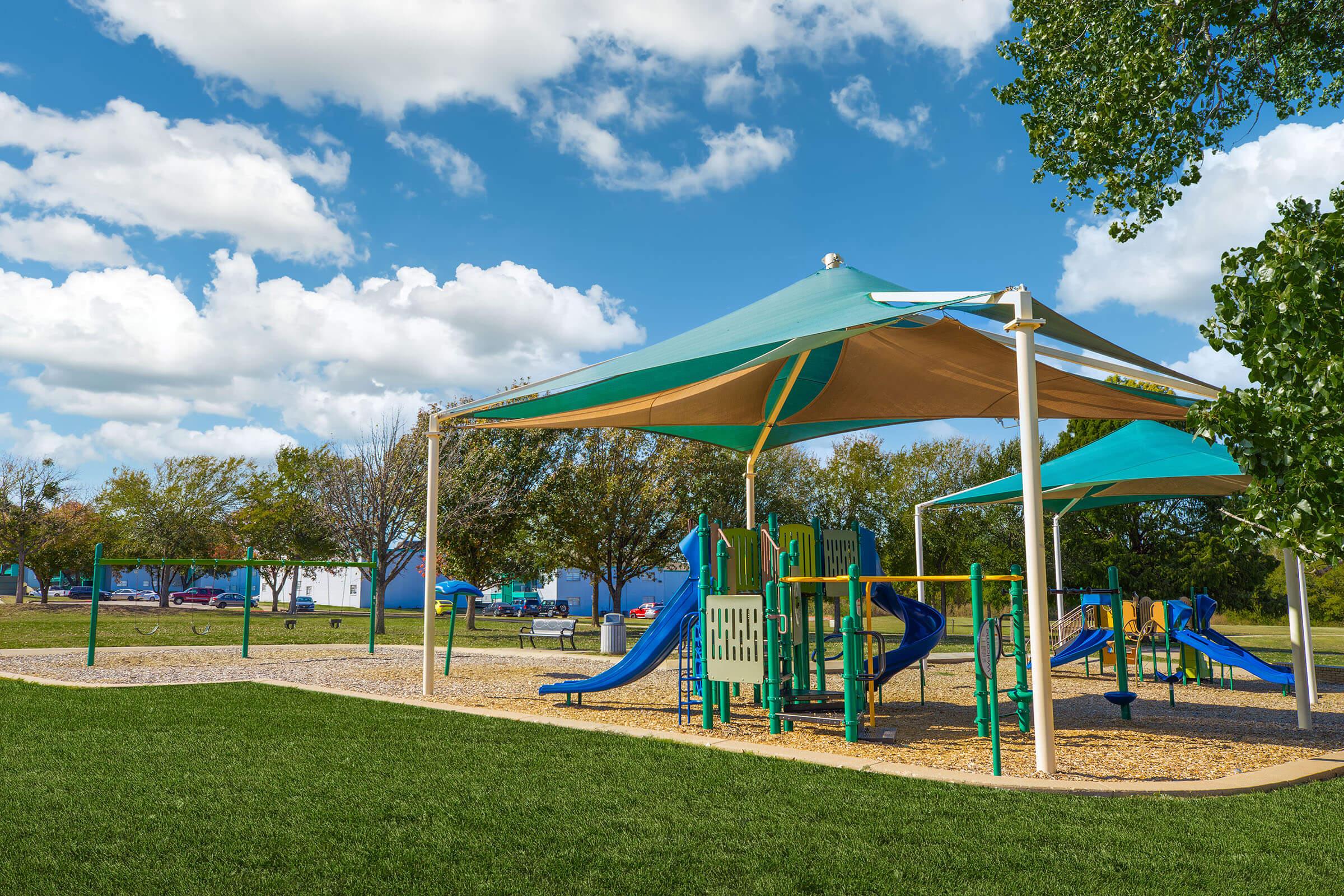 A colorful playground with slides and climbing structures, shaded by large canopies, set in a grassy park. In the background, there are trees and a clear blue sky with fluffy white clouds, creating a vibrant and inviting outdoor space for children to play.