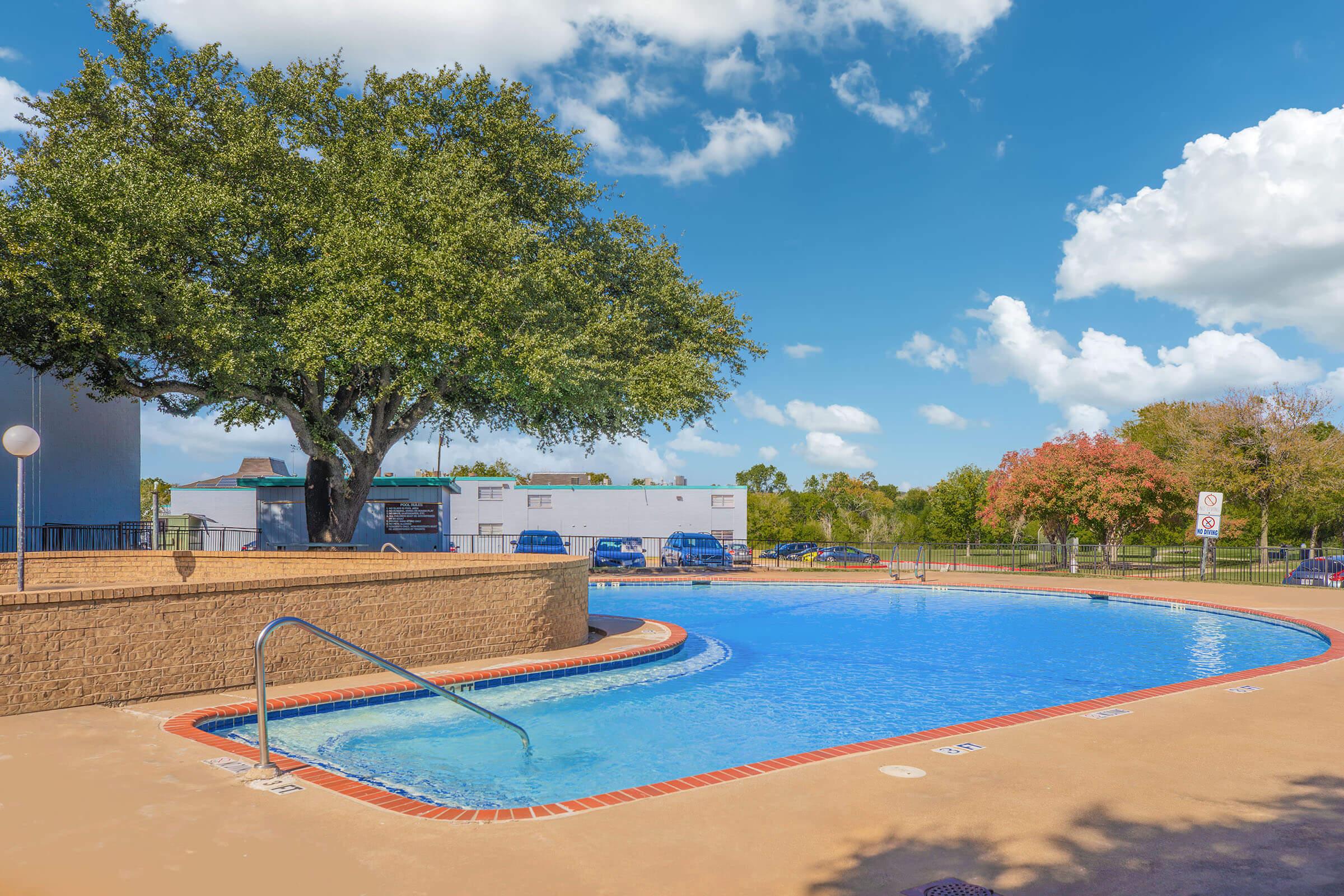 A sunny pool area featuring a clear blue swimming pool surrounded by a tan deck. Lush green trees provide shade nearby, and colorful trees with autumn leaves are visible in the background. A few cars are parked in the distance under a bright blue sky with fluffy white clouds.