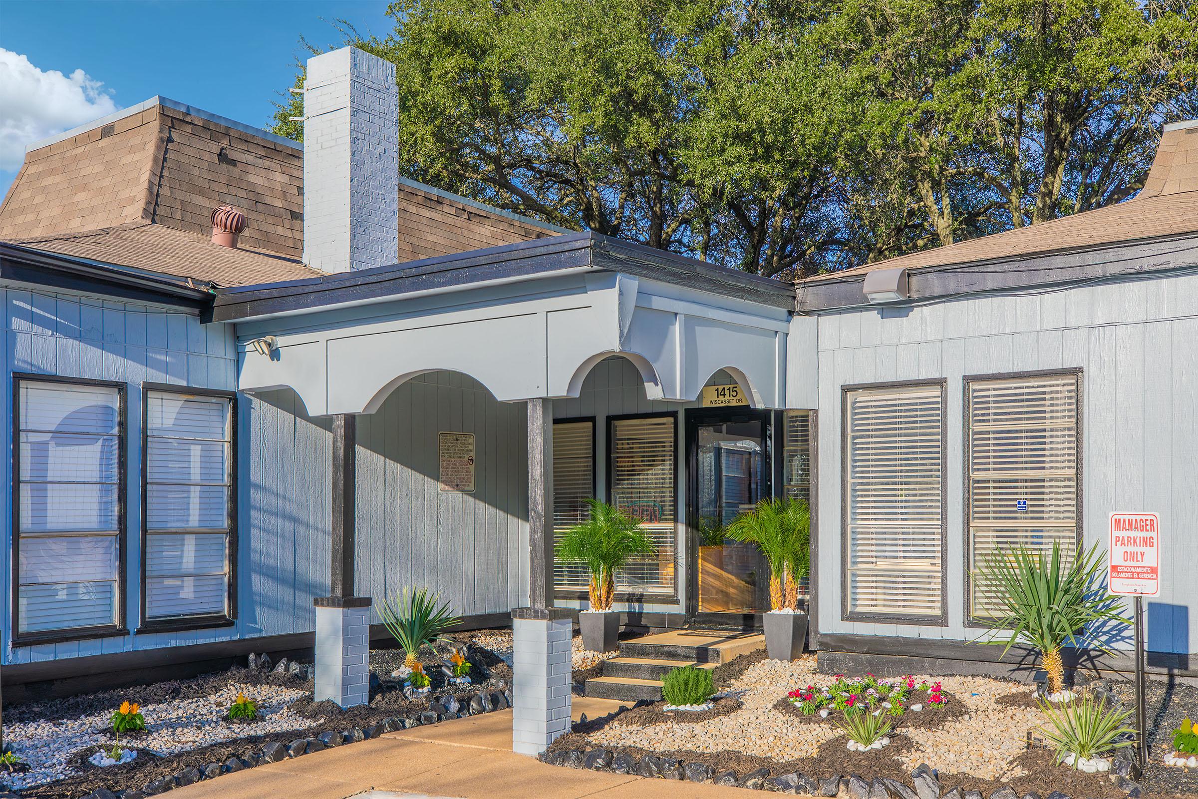 A modern building facade with a gray exterior and a distinct entryway featuring an arched overhang. Lush palm plants and colorful flower beds adorn the front landscape, with light gravel surrounding the entrance. Windows are framed with white shutters, and a warning sign is visible nearby.