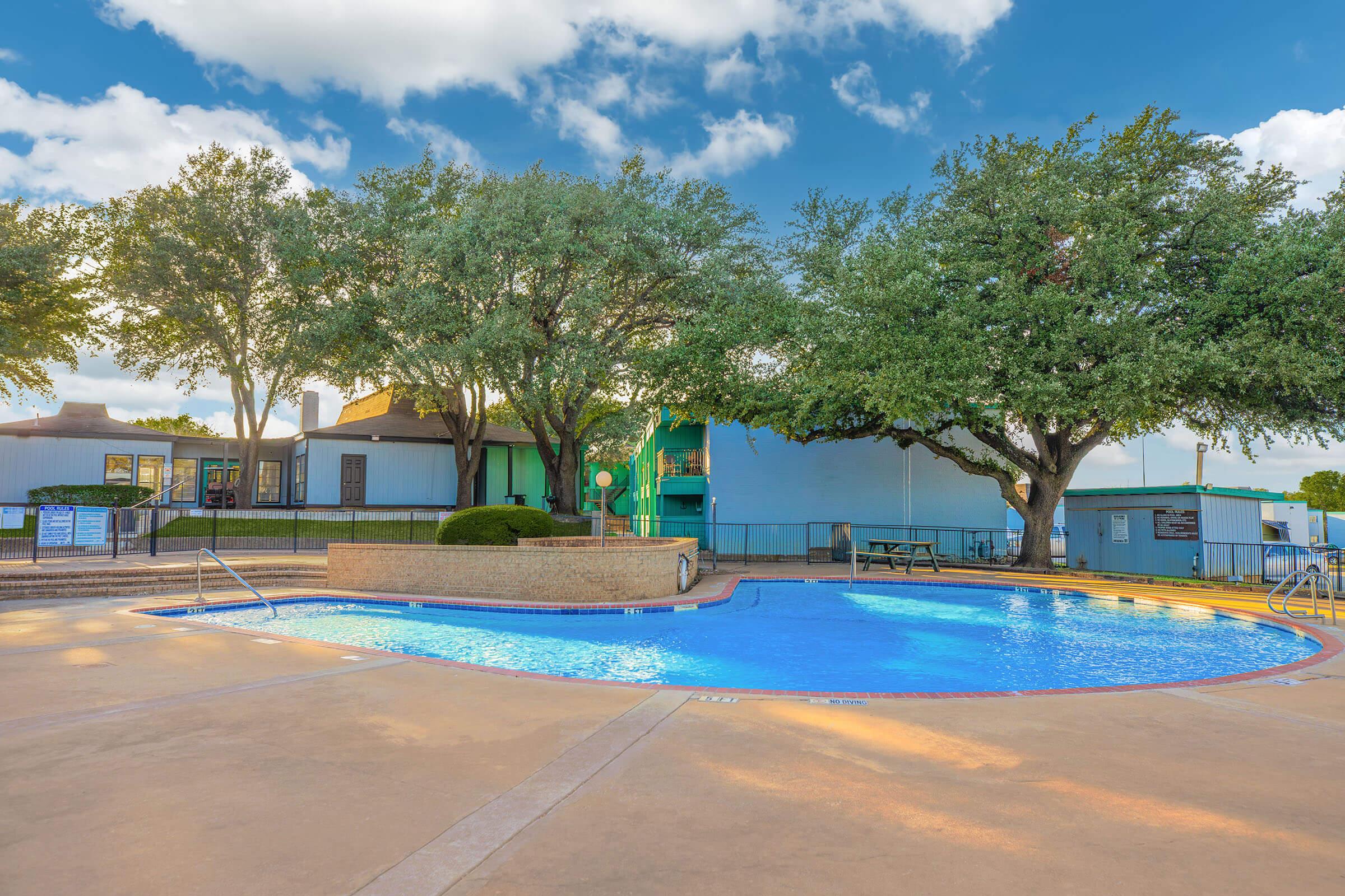A clear blue swimming pool surrounded by concrete, with trees providing shade nearby. In the background, a building features large windows and green accents. The sky is partly cloudy, creating a bright and inviting atmosphere. Picnic tables are visible near the pool area.