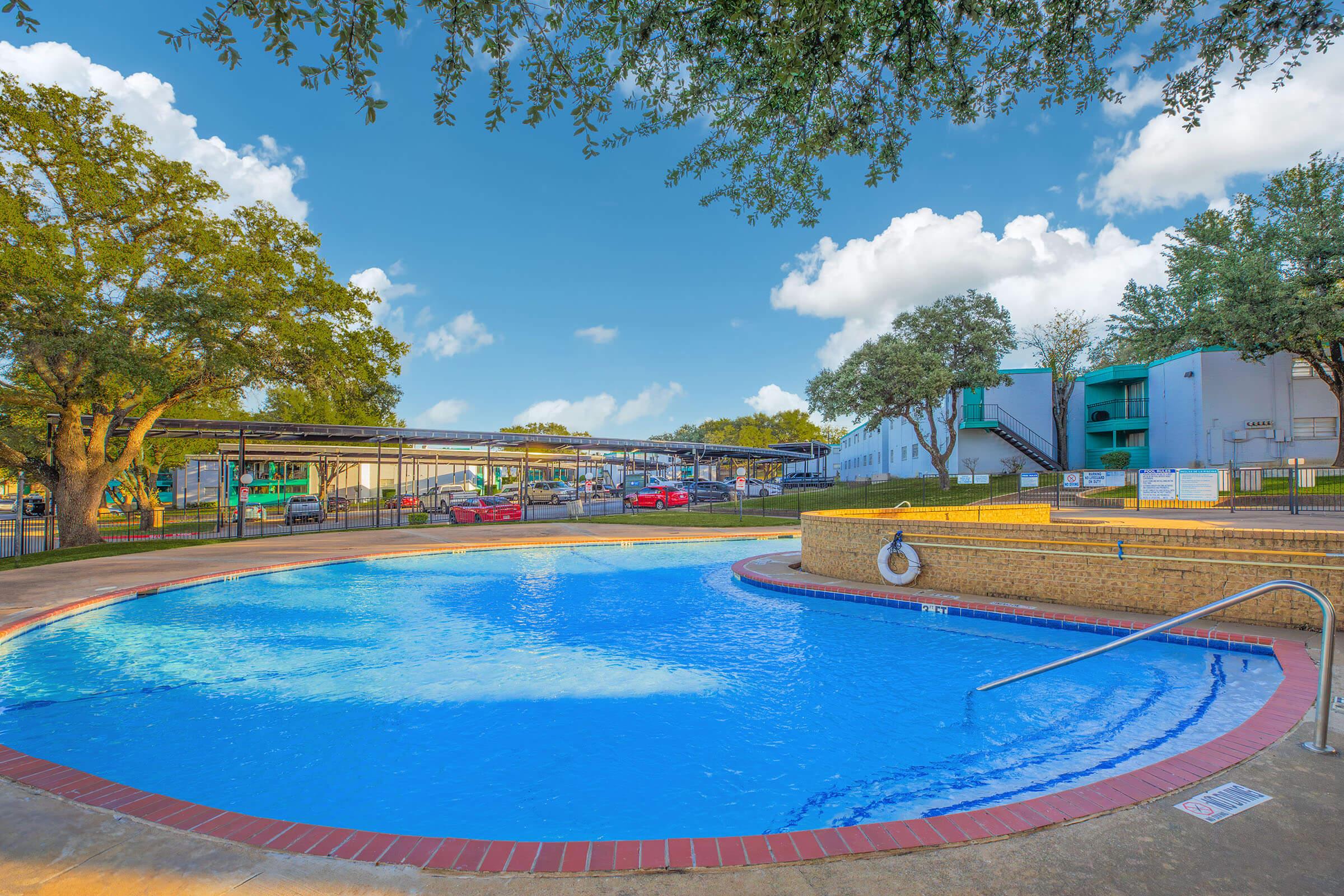A clear, inviting swimming pool surrounded by a paved area and green trees. The pool features a gradual entry with a railing, and in the background, there are buildings with balconies. The sky is bright with fluffy clouds, creating a relaxed, sunny atmosphere.