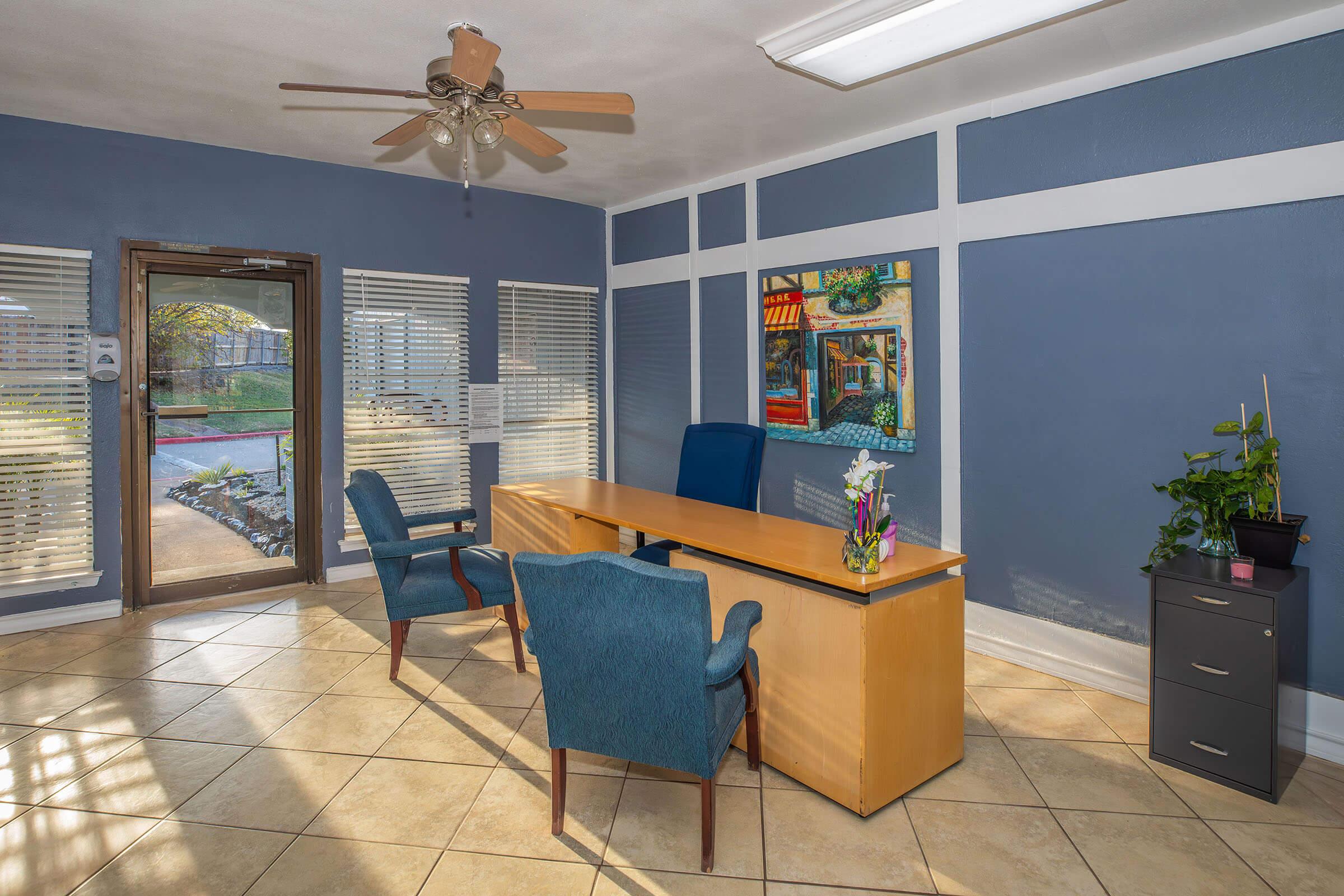 A modern office space featuring a wooden desk, two blue chairs, a potted plant, and a filing cabinet. The room has light-colored tiled flooring, large windows with wooden blinds, and blue walls. Natural light brightens the area, creating a welcoming atmosphere.
