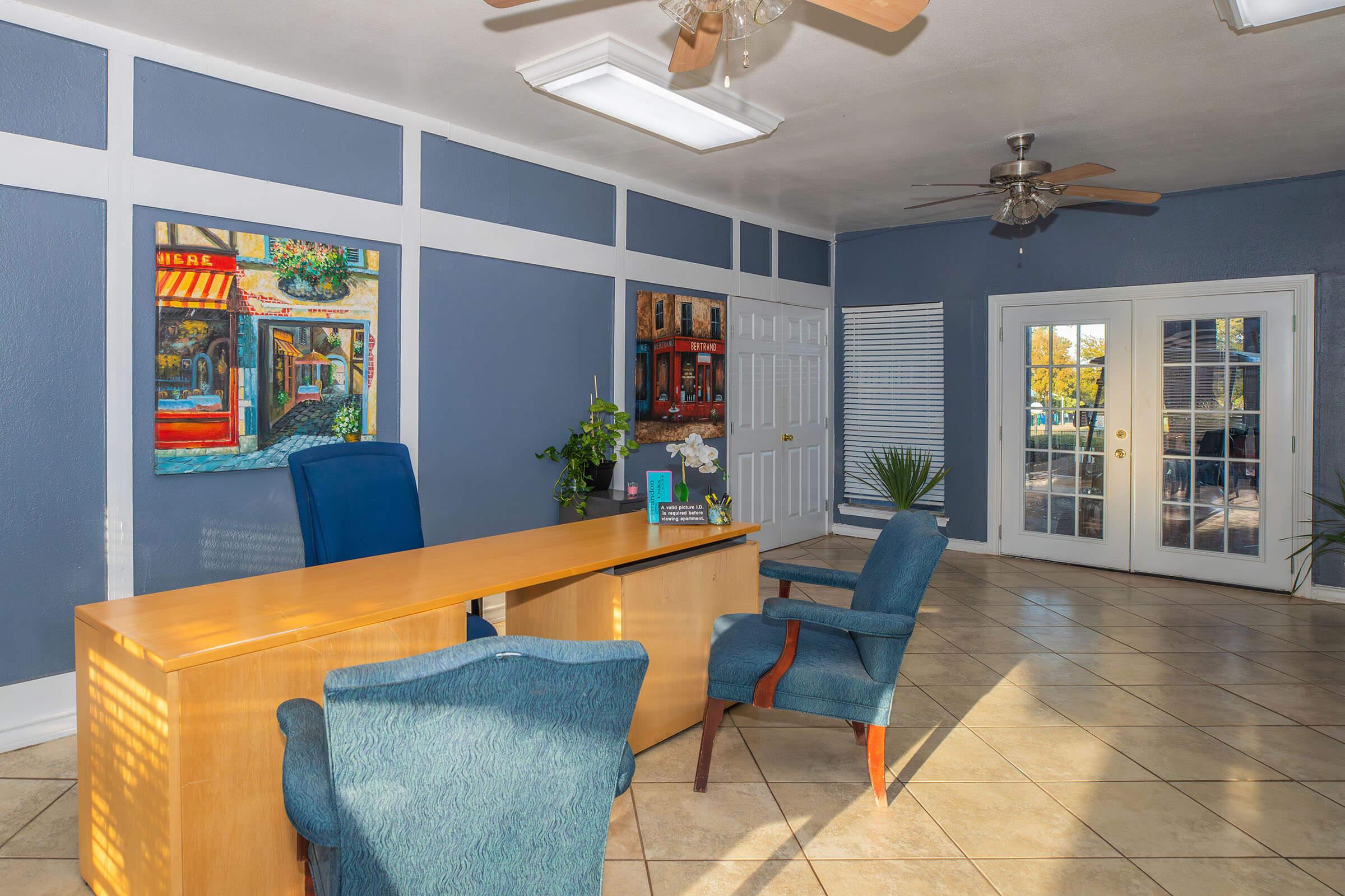A bright and spacious office reception area featuring a wooden desk, two blue chairs, and a colorful painting on the wall. Large windows provide natural light, and there are plants enhancing the ambiance. The walls are painted in a calming blue color, and French doors lead outside.