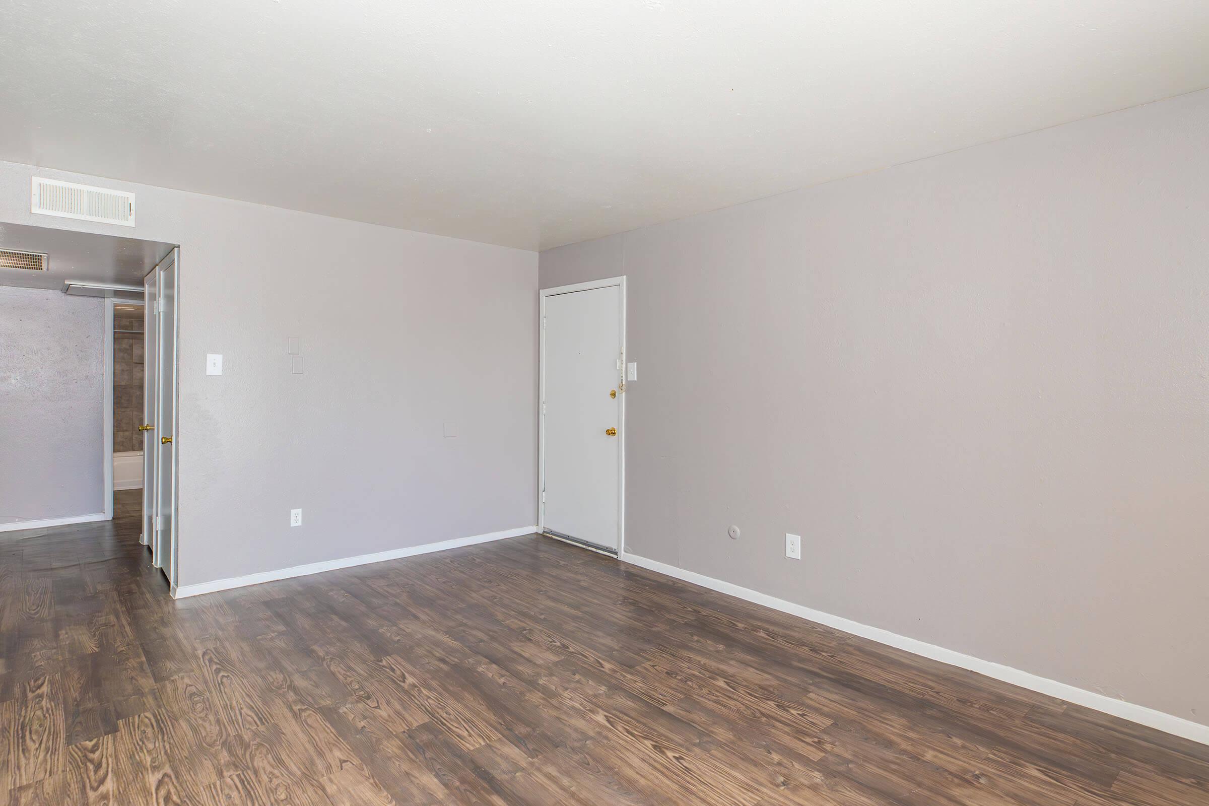 A spacious, empty room with light gray walls and a dark wood laminate floor. There is a plain door on the right side leading outside and an open doorway on the left side. The light source is coming from above, illuminating the space, which has no furniture or decorations.