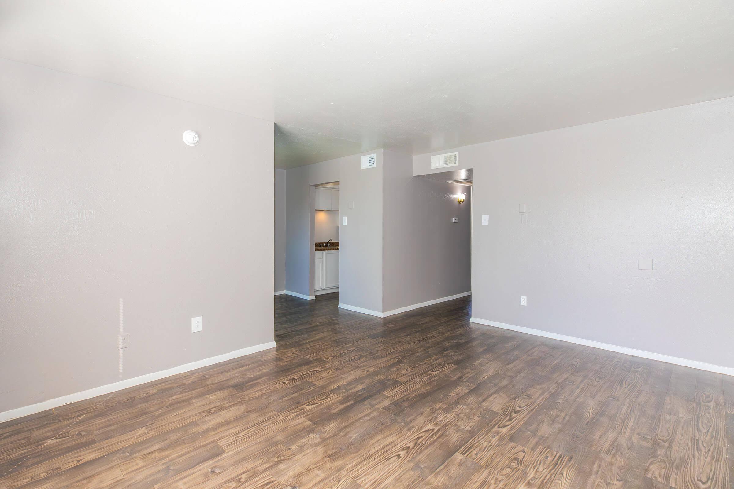 A spacious, empty room with light gray walls and glossy wooden flooring. The room leads to another area with a view of a kitchen in the background. Natural light brightens the space, creating an airy atmosphere. Ideal for personal decor and furniture arrangement.