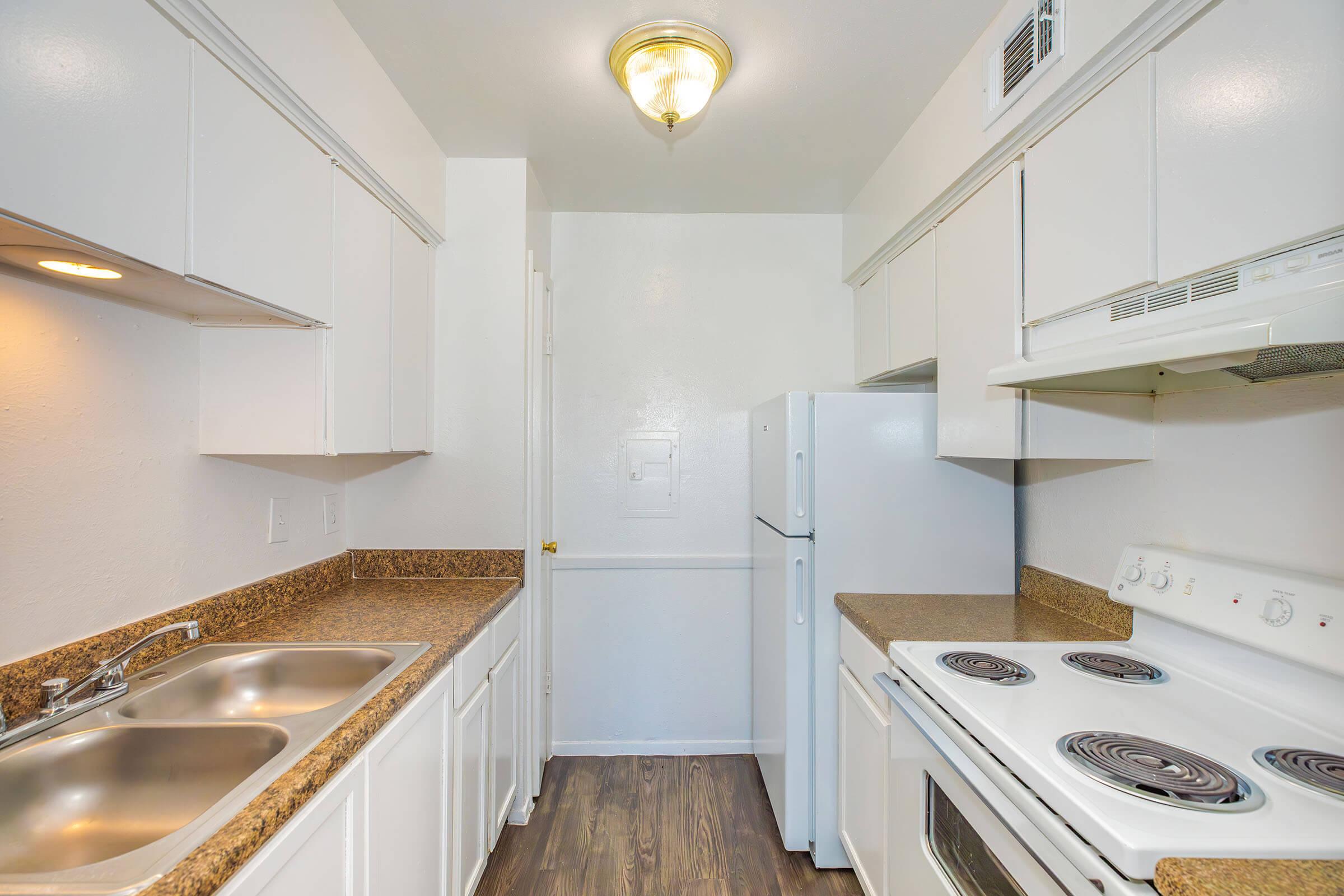A bright, modern kitchen featuring white cabinetry, a double sink, a white refrigerator, and a white stove. The countertops are a light brown granite, and the floor is a dark wood laminate. The space is well-lit with overhead lighting and has a clean, open layout.