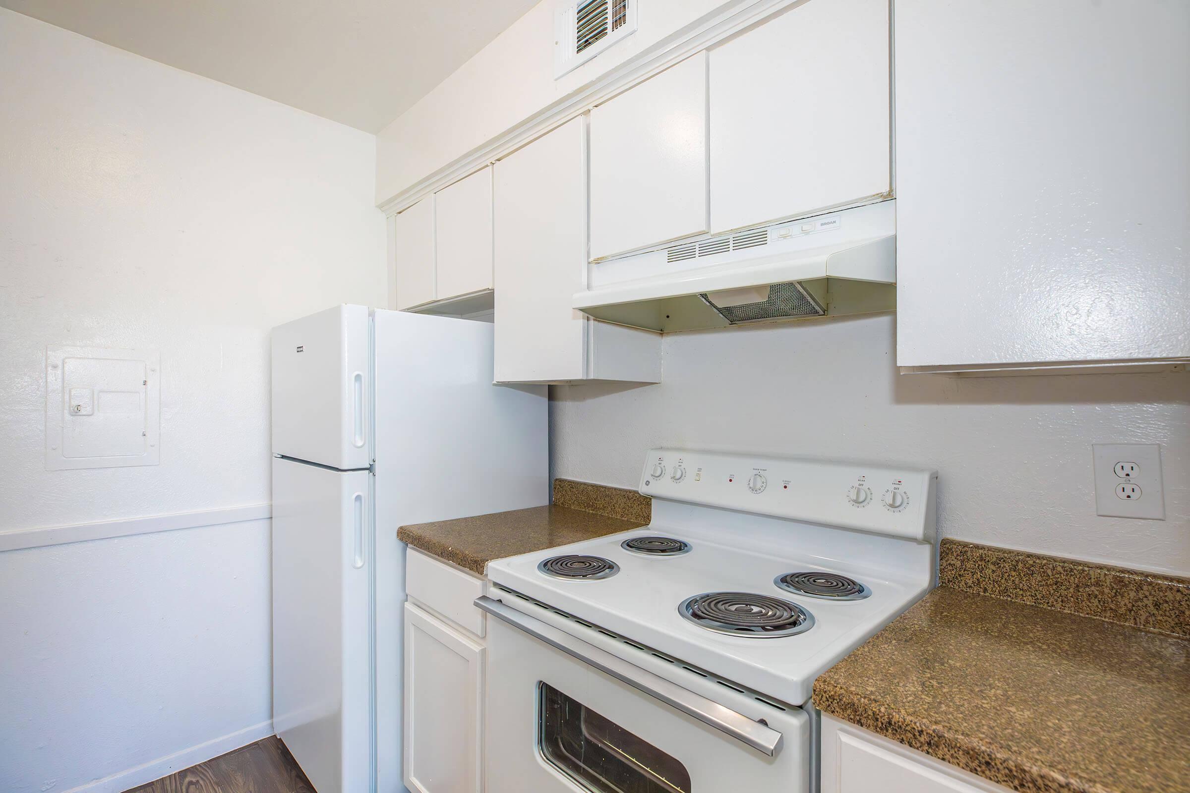 A clean, modern kitchen featuring a white refrigerator, an electric stove with four burners, a built-in microwave above the stove, and light-colored cabinets. The countertops are a dark granite, providing a contrast to the white cabinetry. The walls are painted in a light color, enhancing the bright and open feel of the space.