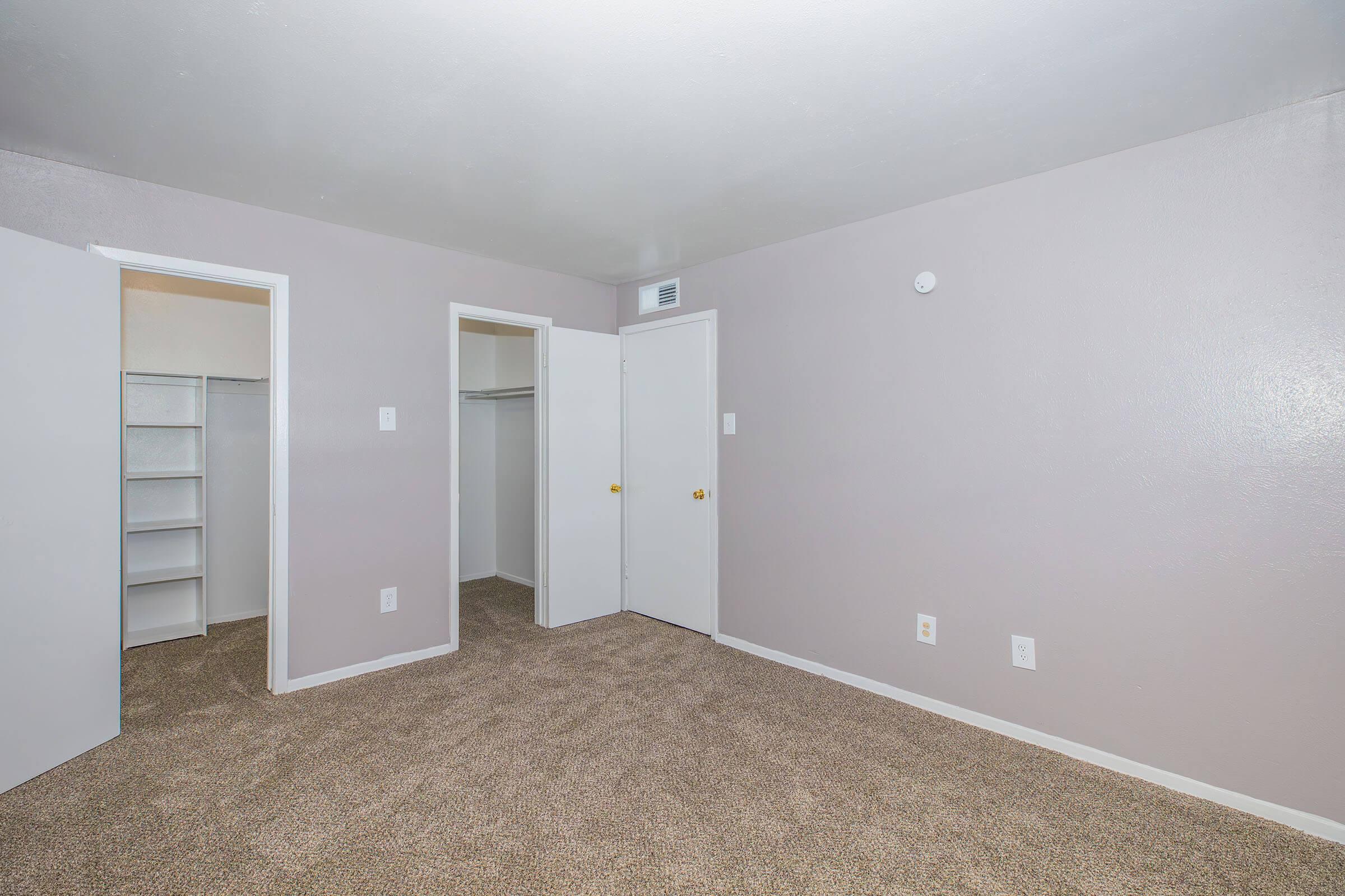 Empty room with light gray walls and carpeted flooring. Two doors on the left lead to closets, one with shelves. A single door on the right, and a small white wall-mounted fixture near the ceiling. The overall atmosphere is clean and spacious, suitable for various interior design choices.