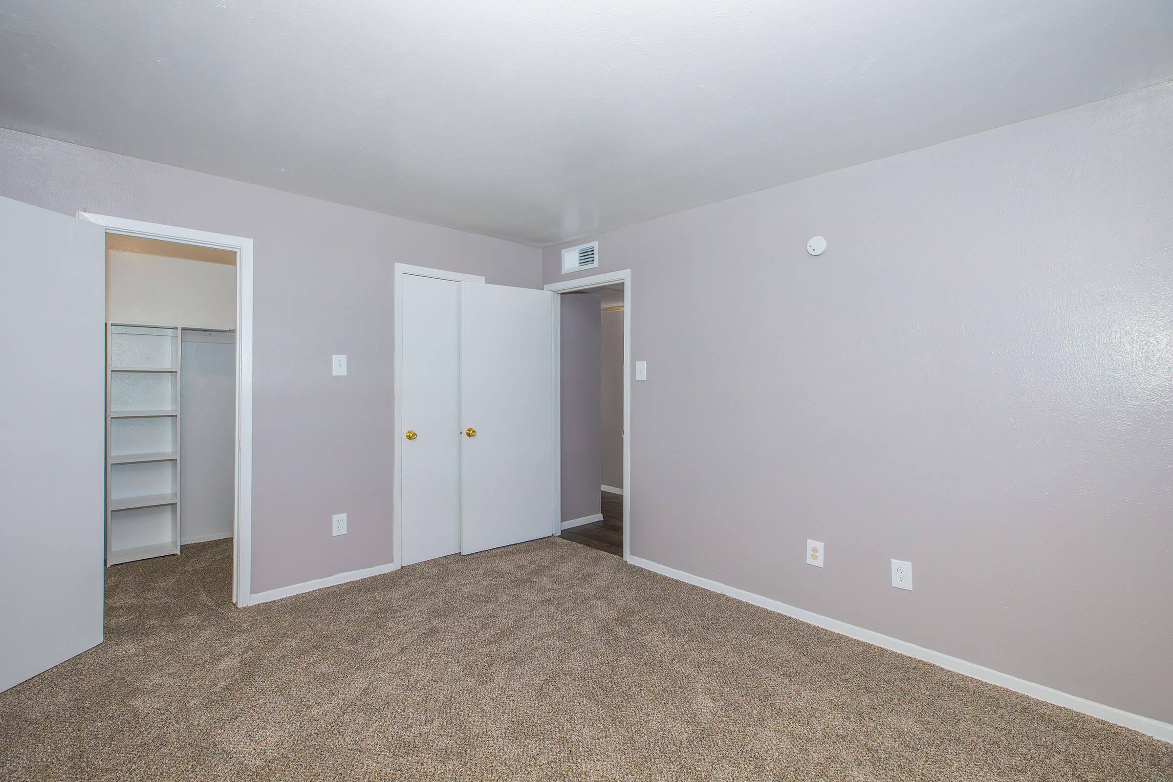 Empty room featuring light gray walls and beige carpet. Two doors are visible, one leading to a small closet with shelves. Natural light comes in, giving a spacious feel. The room has a neutral color scheme and minimal decorations, creating a blank canvas for personal styling.