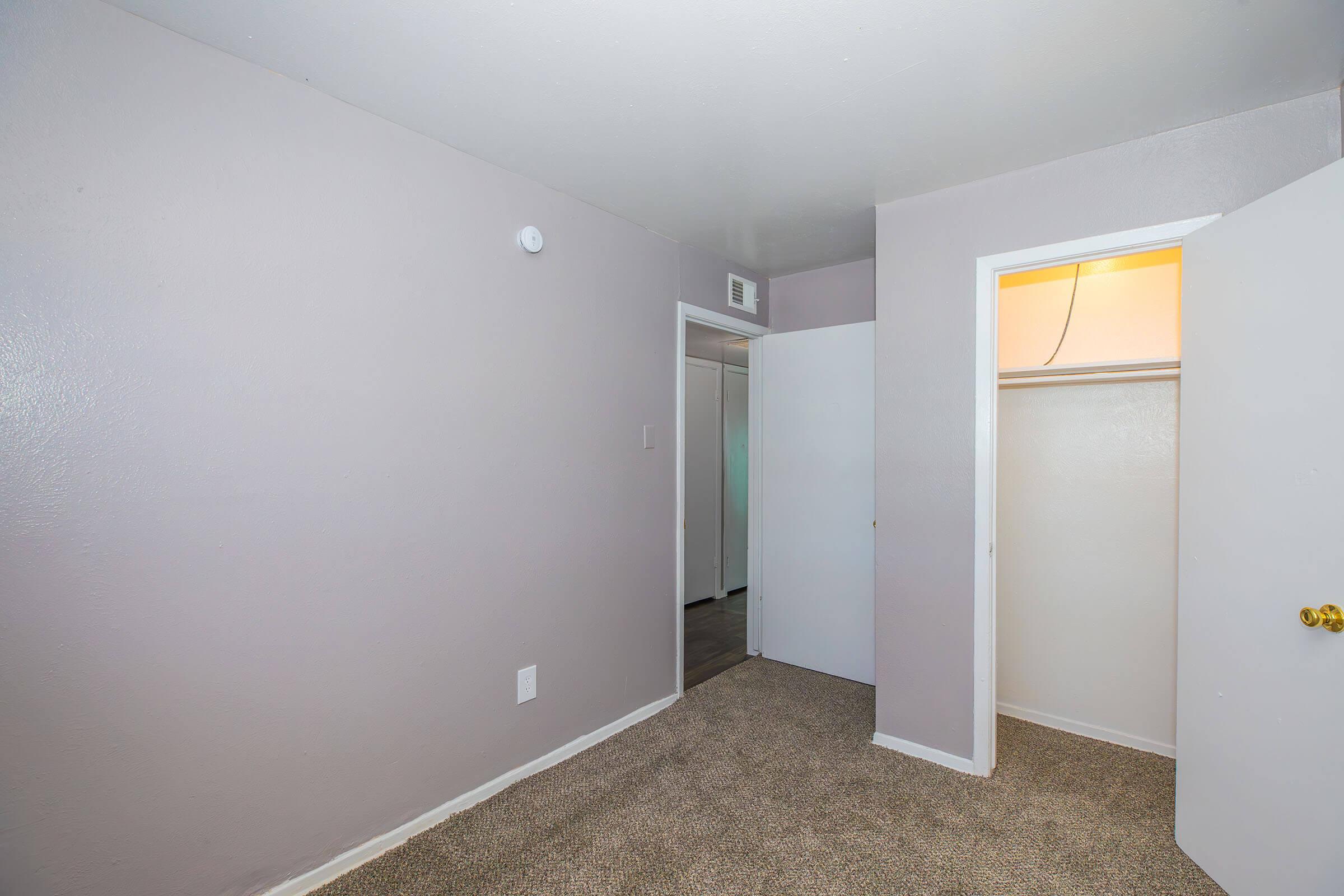 A small, empty room with light gray walls and carpeted flooring. There is a closed door on the right leading to a closet, and another door visible in the background. The room has a simple, uncluttered appearance with a light source coming from the door on the left.
